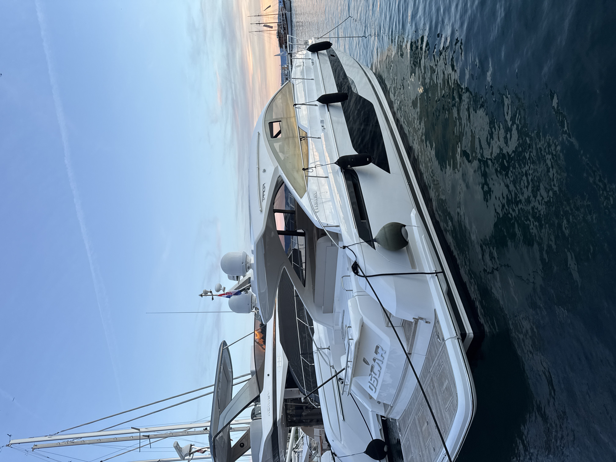 The image captures a serene evening scene at the harbor in Makarska, Croatia. In the foreground, a sleek white motor yacht named "OSCAR" is moored, its polished hull reflecting the twilight sky. The yacht features a dark stripe along its waterline and black fenders. Behind it, another, larger yacht, possibly an Azimut, is partially visible. The sky above is a soft gradient of blue, with hints of pink and orange from the setting sun, and a faint contrail stretches across it. In the background, silhouetted against the horizon, are several other boats, including what appears to be a traditional sailing vessel. The water is calm, with gentle ripples disturbing its dark surface, and the reflections of the boats and the sky are visible. There are no people visible in the image.