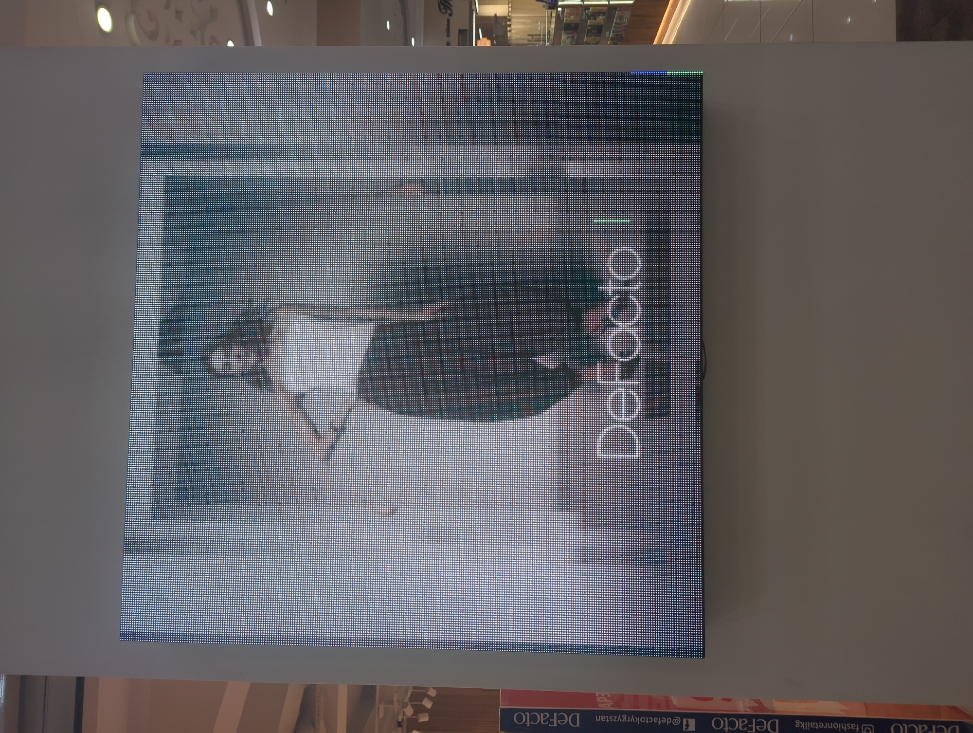 The image shows a large LED display mounted on a wall in what appears to be a shopping mall or commercial space in Bishkek City, Kyrgyzstan. The display features an advertisement for the clothing brand "DeFacto," with a woman modeling an outfit. The background includes glimpses of other shops and the mall's interior. The lighting is bright, suggesting an indoor environment.