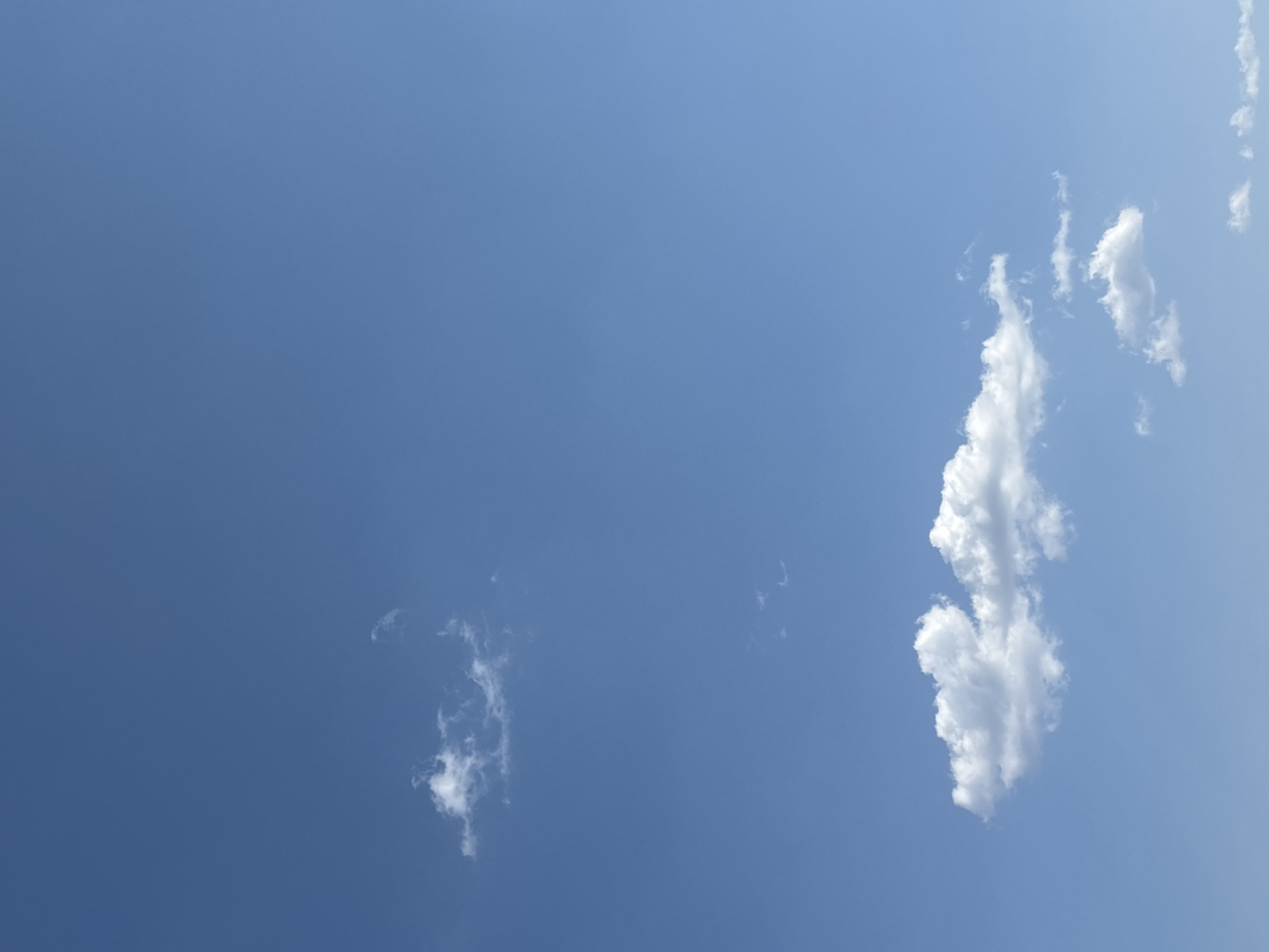 The image displays a clear, light blue sky under daylight conditions, purportedly captured in Santa Clara Ocoyucan, Mexico. Scattered white cumulus cloud formations are visible. A significant, horizontally elongated cloud mass is positioned in the lower-center, exhibiting distinct edges and internal shadows. Above this, in the upper-left quadrant, a smaller, more diffuse cluster of white clouds is present. Additional fragmented white clouds appear near the bottom-right and upper-left edges of the frame. No ground features, artificial structures, or living subjects are visible. The environment consists solely of the atmosphere and cloud formations.