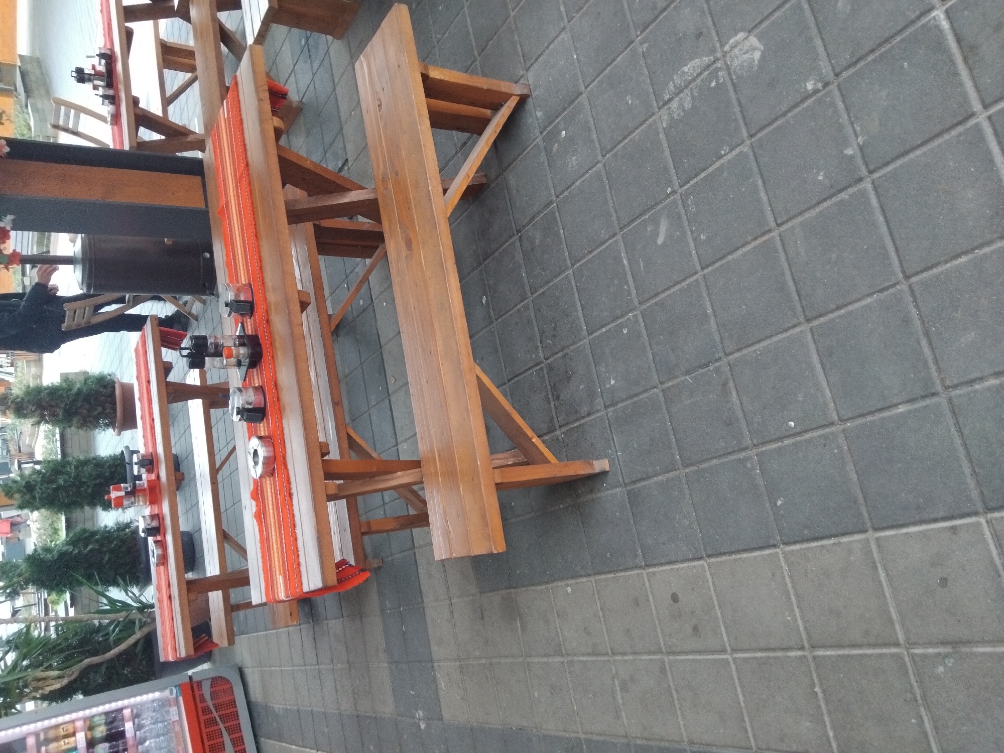 The image shows outdoor seating at a restaurant or cafe. The seating consists of wooden picnic tables and benches with orange and red patterned table runners and condiment sets. A person is seated near a heater. A cooler is visible nearby. Evergreen trees and plants in pots add a decorative element. The ground is paved with gray concrete pavers. The location is likely in the city of Troyan, Bulgaria.