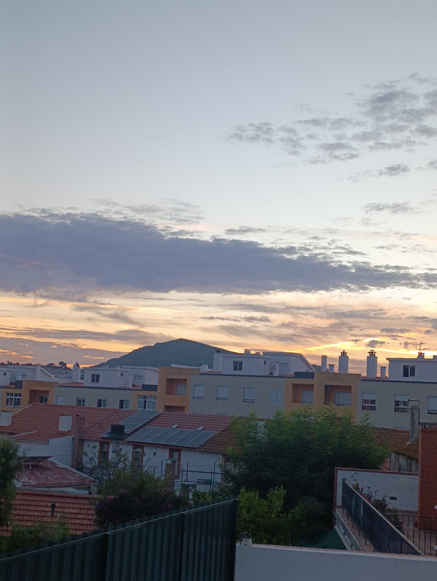 The image depicts a twilight sky over a residential area in Setúbal, Portugal. In the foreground, a dark metal fence partially obscures the view of rooftops with red tiles and solar panels. Several multi-story buildings with light-colored facades and visible chimneys dominate the midground. Beyond the buildings, a green, tree-covered hill rises under a sky streaked with hues of orange, yellow, and purple from the setting sun, with scattered clouds. The overall scene suggests a peaceful, urban evening.