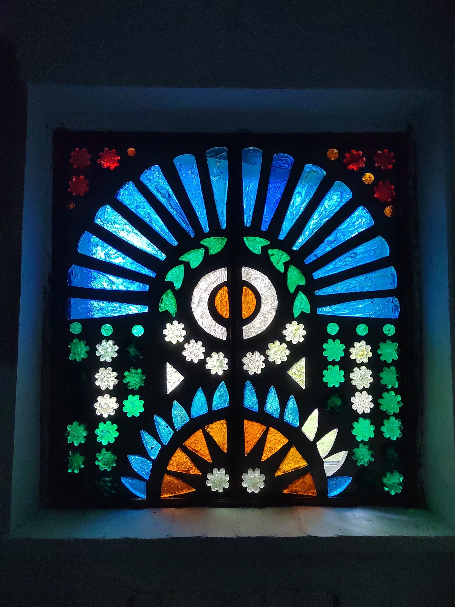 This image shows a square stained glass window, lit from behind. The window is dark in color and contains a variety of colored glass pieces arranged in a symmetrical pattern. The central design resembles an eye or a sunburst, with radiating lines of blue and white glass at the top, and a semicircular arrangement of orange and white glass at the bottom. Scattered around the central design are smaller embellishments of green, white, and red glass in flower and circular shapes. The overall aesthetic is colorful and decorative. The window is set into a dark wall, with a muted gray ledge beneath it. The lighting casts a soft glow on the surrounding wall. The location is noted as Unknown, Unknown.