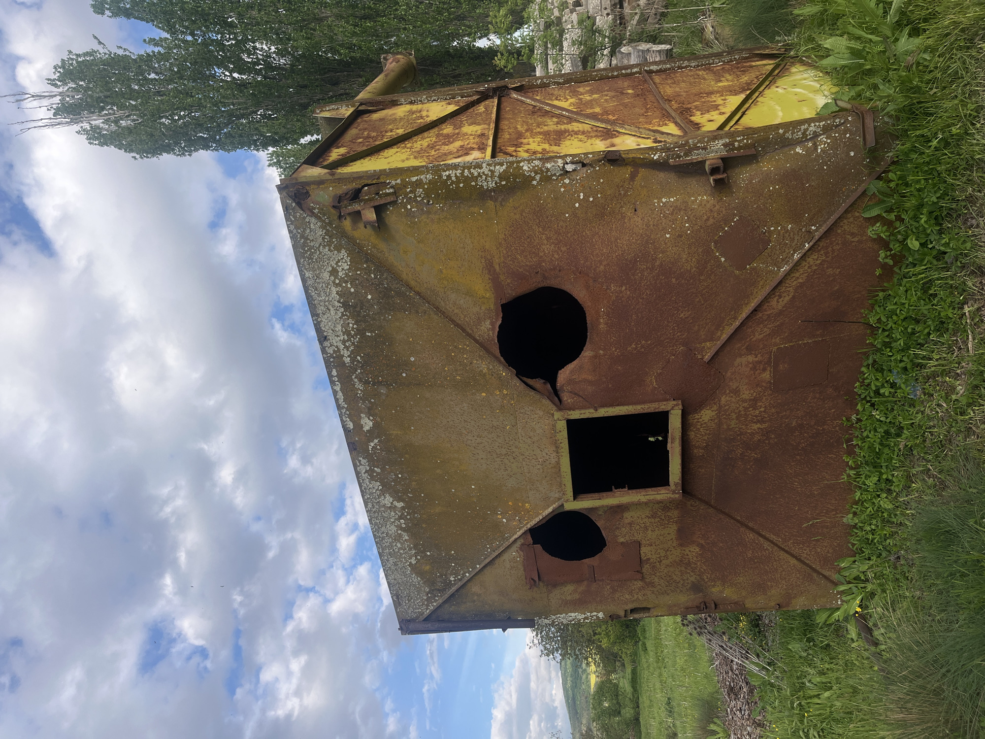 An eye-level, medium shot shows a large, rusty, yellow metal structure with holes in it sitting in a grassy field in Lozarevo, Bulgaria. The sky is visible with white clouds and blue sky. There are trees in the background and weeds are growing around the bottom of the structure. The structure appears to be old and possibly abandoned.