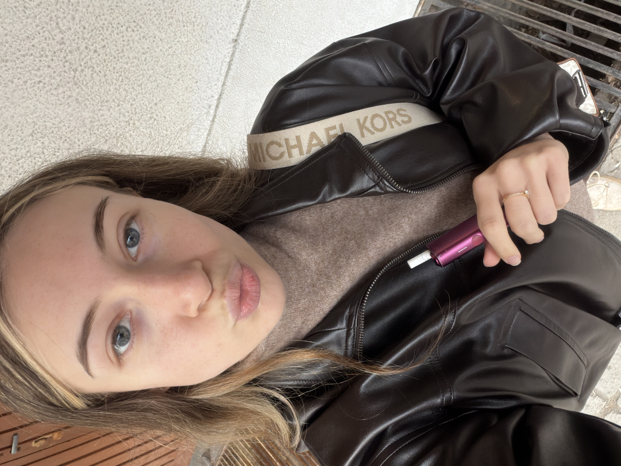 The image shows a selfie taken by a woman outdoors. She has light skin, blue eyes, and light brown hair with blonde highlights. Her lips are pursed as if posing for a kiss. She wears a brown sweater and a black Michael Kors jacket. In her hand, she holds a purple device, possibly an e-cigarette or similar item. The background includes a tiled surface and a metal grate, which might be part of a drain or ventilation system. A wooden bench is visible at the bottom of the image. The location appears to be an urban public space, possibly in Plovdiv, Bulgaria.