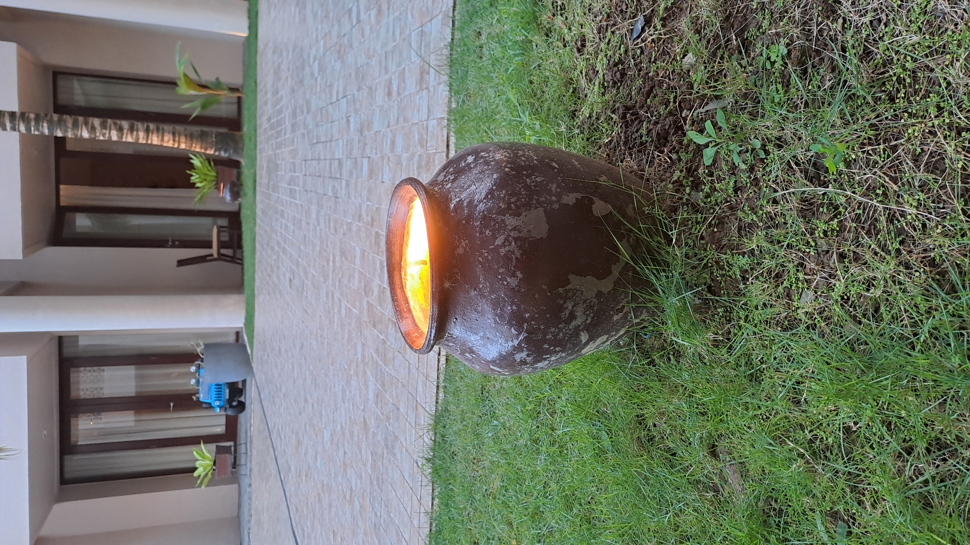 A dark, weathered ceramic pot sits on a patch of green grass. The pot is filled with what appears to be liquid, possibly oil, and a bright yellow flame is burning within it, casting a warm glow. The background shows a paved area with a repeating square tile pattern, leading to a building with large glass doors and white pillars. A small, blue toy jeep is visible in the distance near one of the doorways. The environment suggests an outdoor setting, possibly a courtyard or patio area of a residential or commercial property.