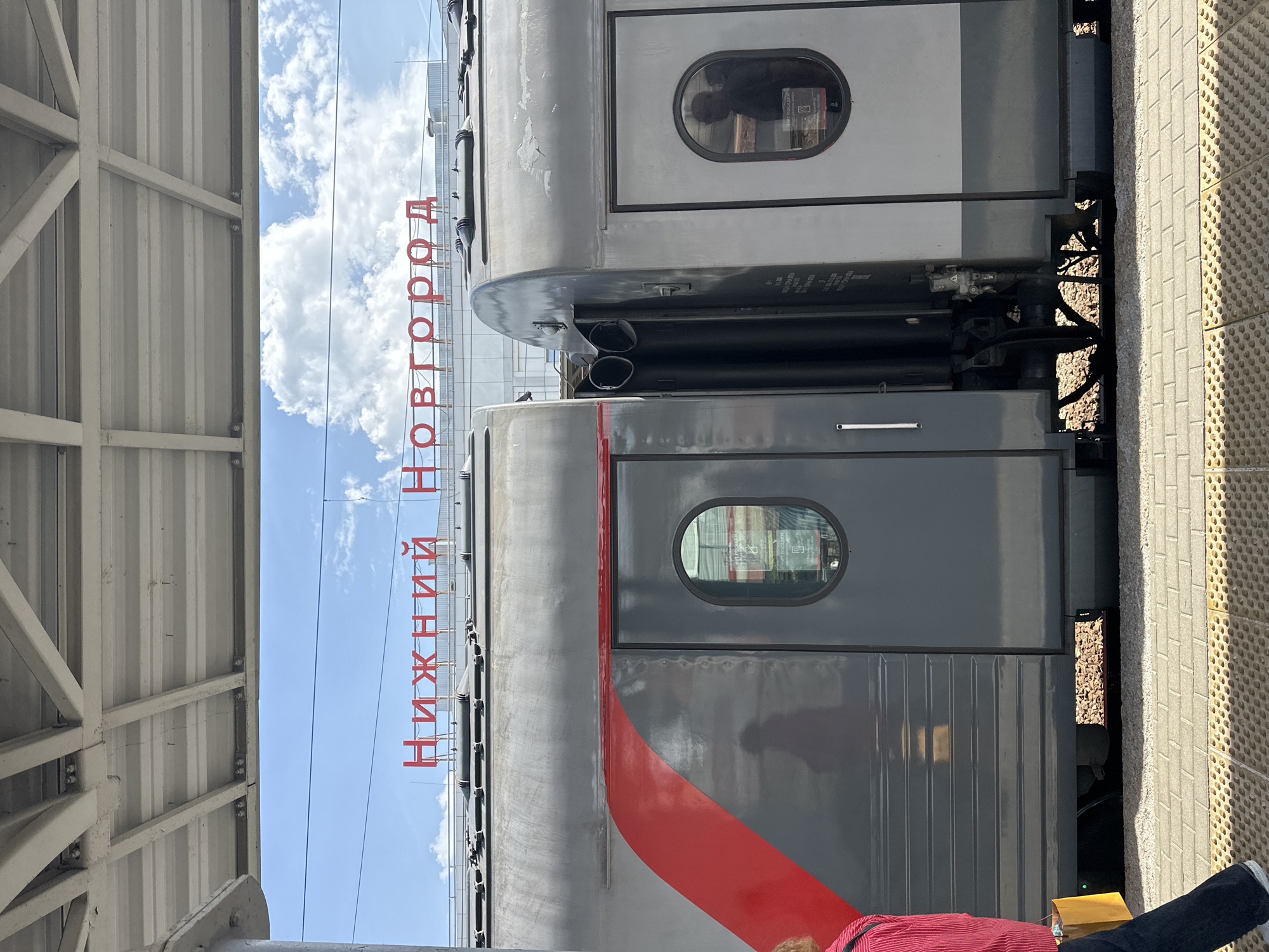 The image shows the side of a train at a station in Nizhny Novgorod, Russia. The red Cyrillic text "НИЖНИЙ НОВГОРОД" is prominently displayed on the side of the train car. The train is gray with red accents. Passengers can be seen reflected in the oval windows of the train. The station platform is visible on the right side of the frame, and a portion of the station's roof structure is seen on the left side. The sky is blue with scattered white clouds. A person wearing red stripes and blue jeans can be seen in the lower right corner of the image. The scene takes place during the day.