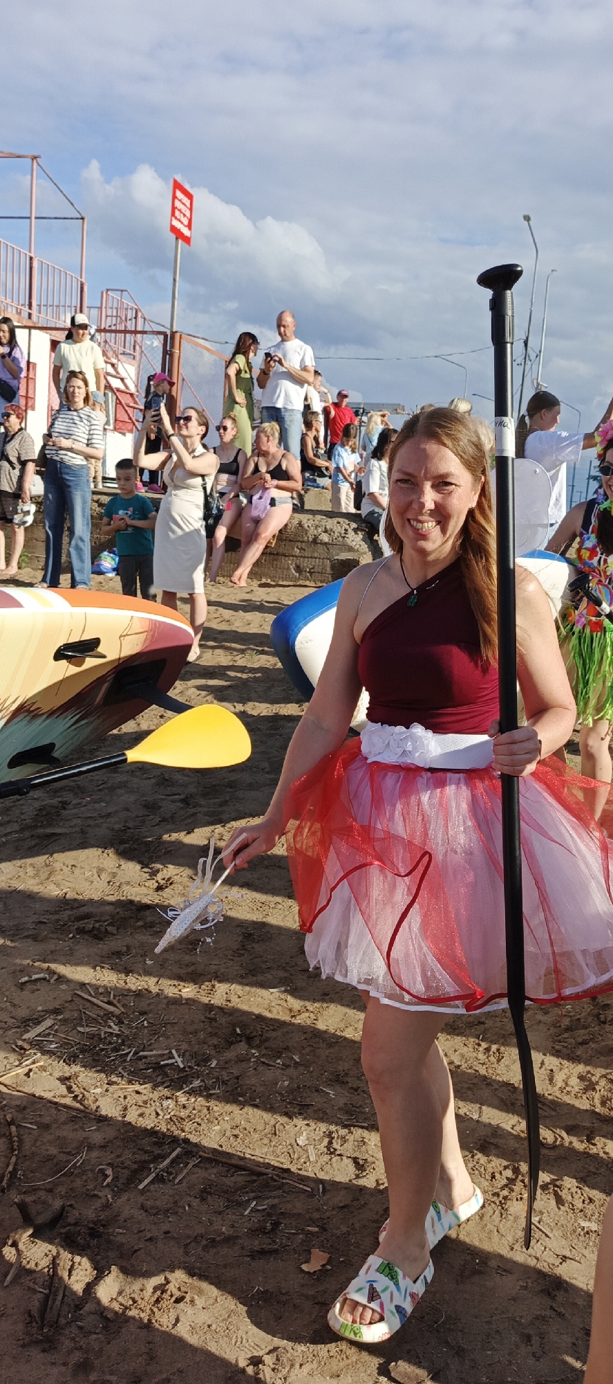 A full-body shot captures a smiling woman standing on a sandy beach, dressed in a red and white tutu and a burgundy top. She holds a paddle and a white feather ornament. Behind her, a crowd of people, some seated and others standing, are visible near a grandstand. Several paddleboards are arranged in the foreground. The scene is illuminated by sunlight, casting shadows on the sand. The sky is cloudy with patches of blue.