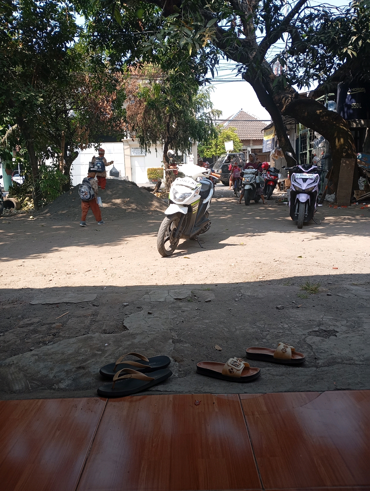 In Ketegan, Indonesia, a sun-drenched dirt road is partially shaded by a large tree. In the foreground, on a tiled surface, a pair of black flip-flops rests beside a pair of tan sandals. The road is occupied by several parked motorcycles, including a white and green scooter, a white scooter, a red scooter, and a light purple scooter. Two children, one wearing a blue backpack and orange pants, and another with a similar backpack and red and white striped shirt, are standing on a pile of dark grey gravel. Behind them, a white building with a sign is visible. The overall scene depicts a typical street in a residential area.