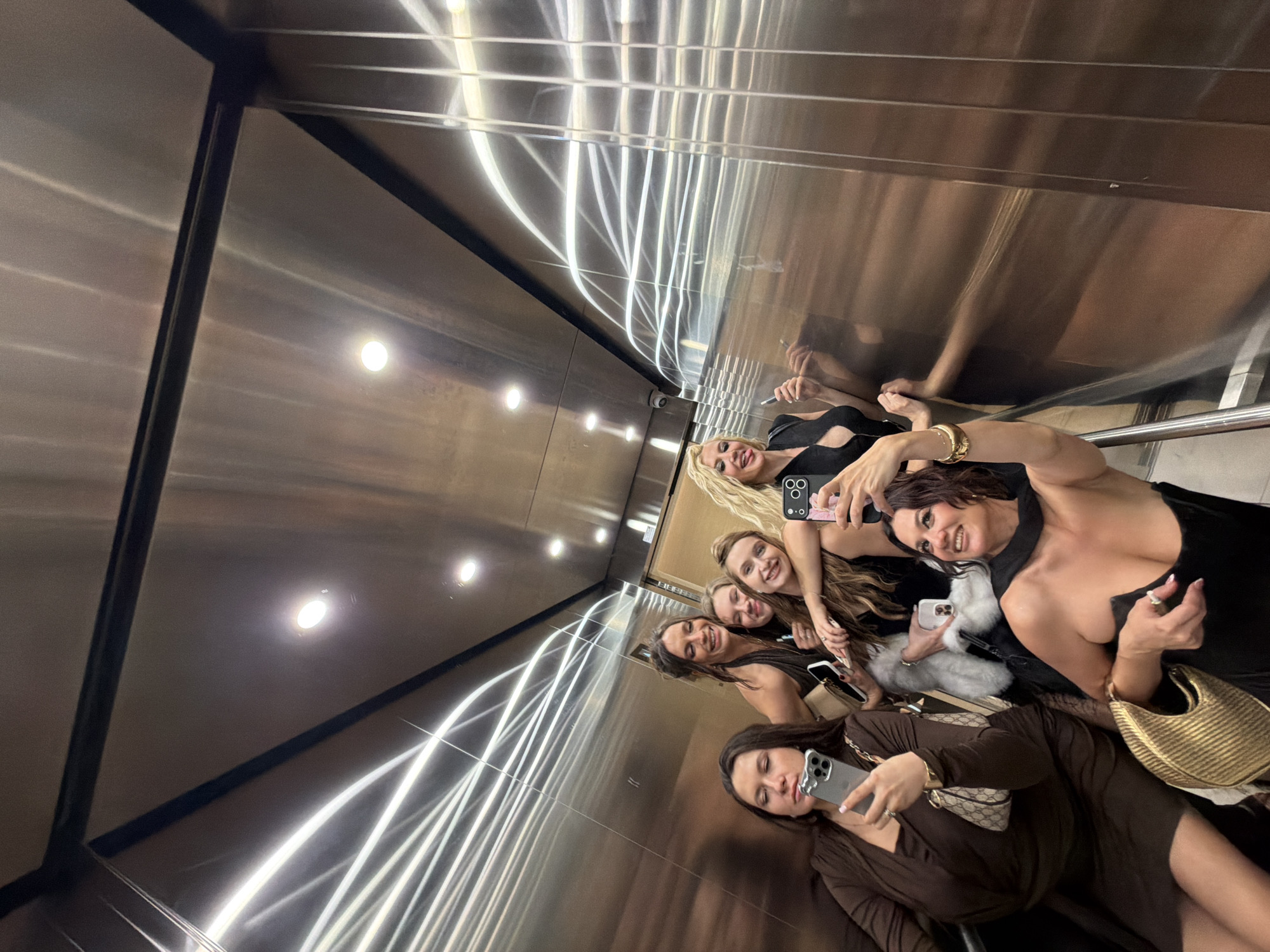 A group of six women is taking a selfie inside a modern elevator. The elevator features stainless steel walls with reflective light fixtures. All the women are dressed in evening attire, including dresses in black and brown tones. They are each holding smartphones to capture the photo. The photo appears to have been taken in Belgrade, Serbia.