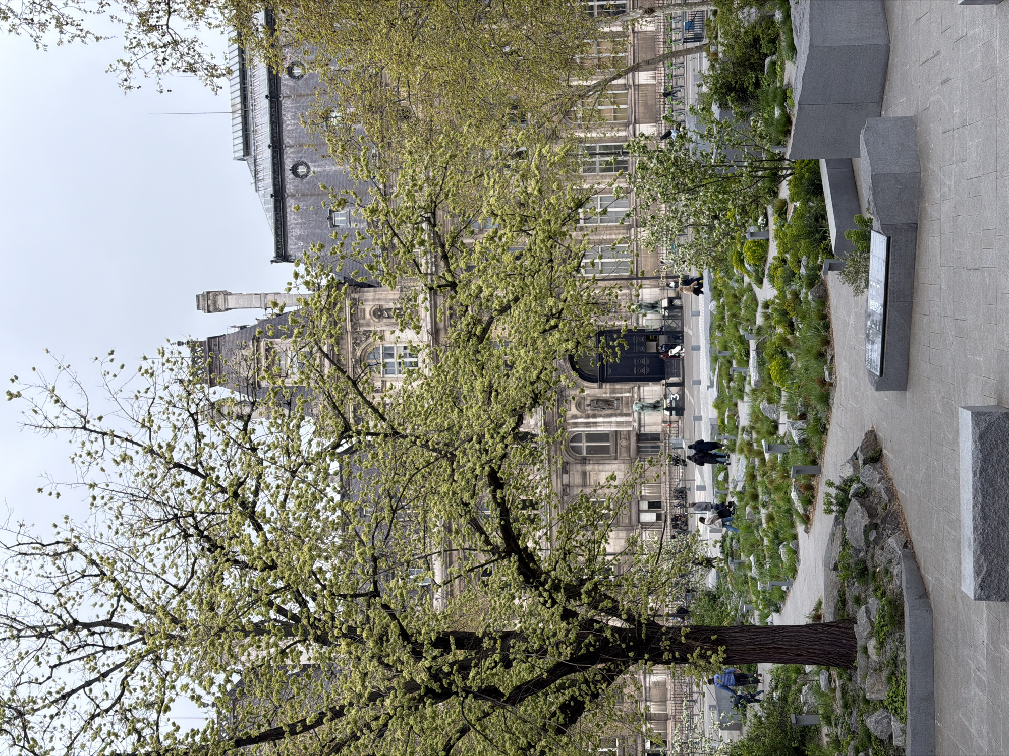 The image depicts a view of the Mémorial des Martyrs de la Déportation in Paris, France. The memorial is visible as a series of low, rectangular markers set in grass and interspersed with stones. A paved pathway runs alongside the memorial. Several people are walking along the path and among the markers. In the background is a large, ornate building, partially obscured by trees with green foliage. The sky is overcast. Stone benches are also visible.