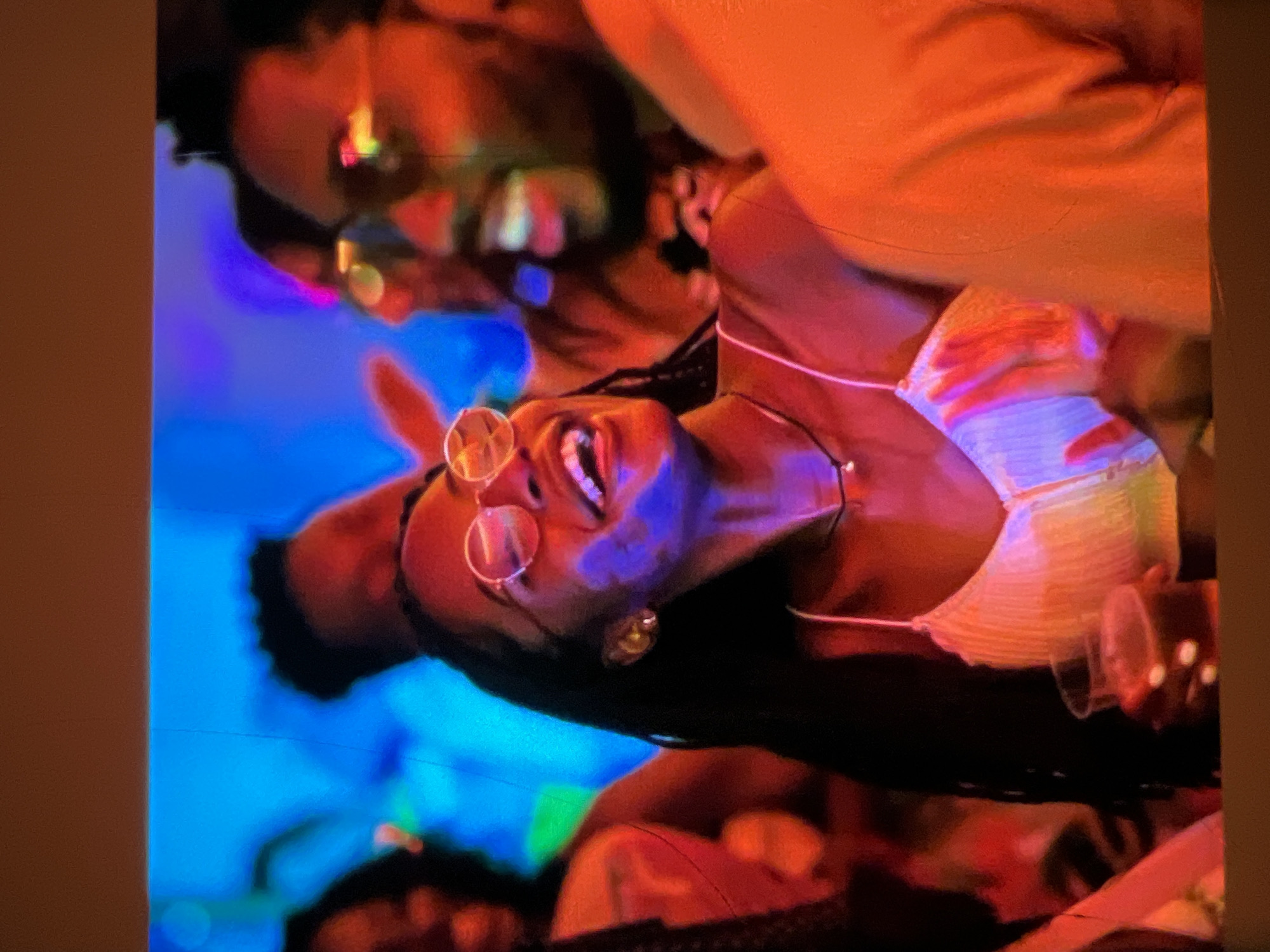 The image captures a lively scene at a nighttime event in Lagos, Nigeria, likely a club or party, characterized by vibrant blue and orange lighting. A young Black woman with long dark hair, wearing round, clear-rimmed glasses, a white camisole, and a thin necklace, is the central focus. She is shown laughing or smiling broadly, with her head tilted back. Her left hand is holding a clear plastic cup containing a dark liquid, possibly a beverage. To her right, another individual, a Black man wearing glasses with reflective lenses, is partially visible, also appearing to be smiling or engaged in conversation. Other blurred figures and colorful lights suggest a crowded and energetic atmosphere. The overall impression is one of social gathering and enjoyment.