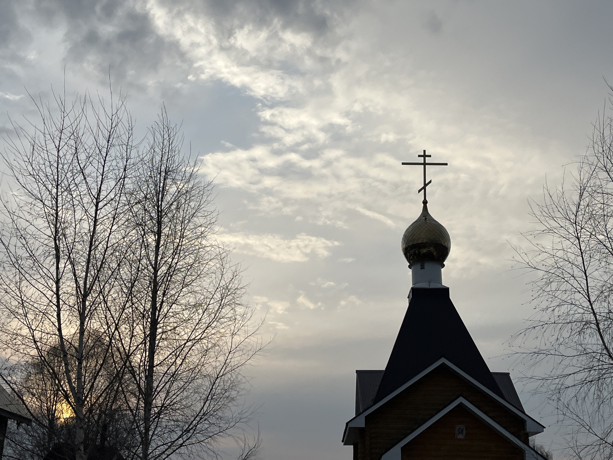 The image shows the top of a wooden structure, presumably a church, with a golden dome and a cross atop it. The architecture suggests it is located in Павловка, Russia. Two bare trees are on either side of the building. The sky is overcast with many clouds. The sun appears to be setting behind the trees on the left.
