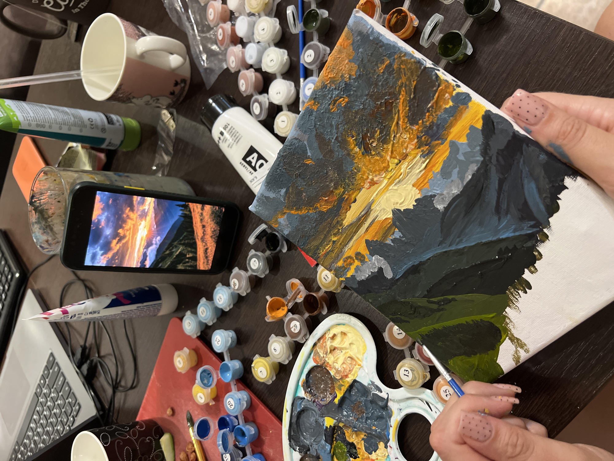 The image shows a person engaged in a paint-by-numbers project on a dark-colored table. The painting depicts a landscape, possibly mountains at sunset, rendered in a variety of colors from dark blues and greens to yellows and oranges. The person is holding a paintbrush in their right hand, applying green paint to the lower section of the canvas.
Around the painting and the person's hand are various art supplies including a plastic paint palette with mixed paints, numerous small containers of numbered paint colors, and a bottle of white acrylic paint labeled "ACRYL/AC". A phone displaying a landscape with a sunset serves as a reference image. Other objects on the table include a mug, a tube of cream, a laptop and various other painting tools.
The setting suggests a home or studio environment, judging by the relaxed arrangement of personal items and art supplies. There's no specific evidence to suggest the location is Podgorica, Montenegro.