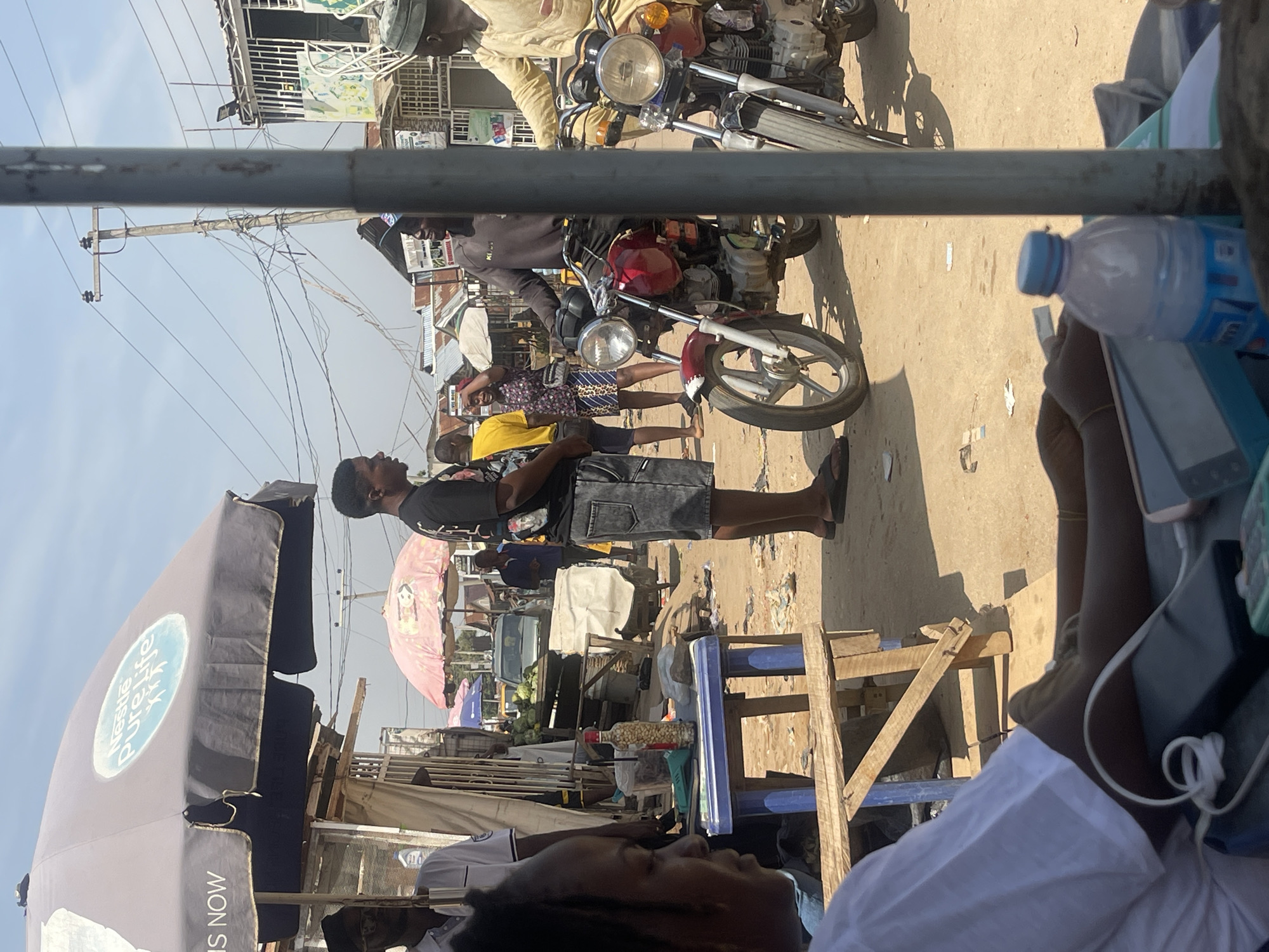 This image captures a street scene in Abuja, Nigeria, likely a marketplace or busy intersection. In the foreground, a person is partially visible from the side, wearing a white t-shirt and appearing to be interacting with a mobile device and a power bank on a table. To the left, a large grey umbrella with the "Nestle Pure Life" logo is prominently displayed.
Behind the foreground subject, the scene opens up to a bustling environment. Several people are visible, some standing and others on motorcycles. Two motorcycles are parked in the center of the frame, one red and one white. A man in a black t-shirt and denim shorts is standing near the red motorcycle, and another man in a yellow top is standing to his left. Further back, more individuals and market stalls with umbrellas are visible, along with a parked car. The ground is dusty and appears littered with some debris. Power lines crisscross overhead, indicating an urban setting. The sky is bright and appears to be a clear day.