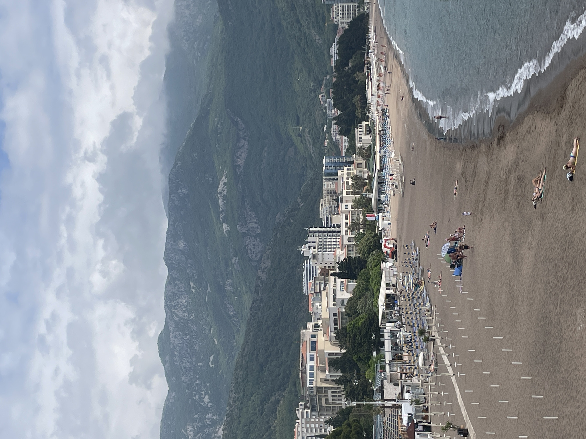 The image captures a coastal scene in Bečići, Montenegro. The photograph presents a vertical panorama where the frame is divided between the sea, a sandy beach, and a mountain range. The beach is populated with beachgoers, some swimming in the sea, others relaxing on the sand. Sunbeds and sun umbrellas are neatly arranged on the beach. Adjacent to the beach is a road, which is also lined with objects resembling sunbeds or loungers. The mountain range, covered in lush greenery, forms the backdrop. The sky overhead is overcast with heavy cloud cover. The buildings of Bečići are visible, nestled between the beach and the mountain, giving a sense of the town's layout and proximity to the natural landscape.