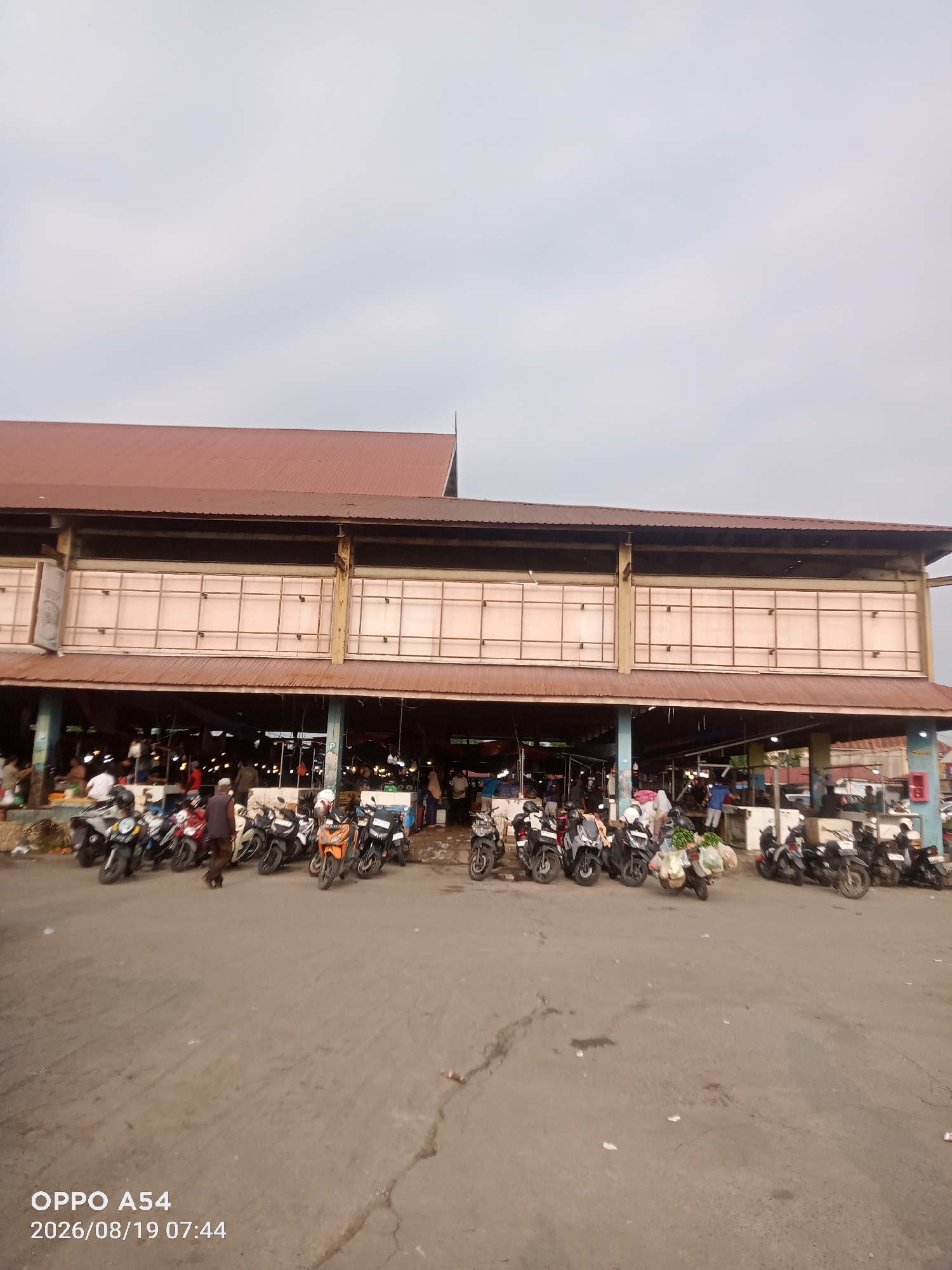 The image depicts an outdoor market in Jambi, Indonesia, on a cloudy day. The market appears to be a covered structure with a reddish-brown roof and beige panels on the upper facade. Stalls are visible underneath the covered area, with numerous motorcycles parked in front of and around the market. People are present, with some individuals walking and others appearing to be vendors or shoppers. The ground in front of the market is paved asphalt with some cracks. In the foreground, a watermark reads "OPPO A54 2026/08/19 07:44."