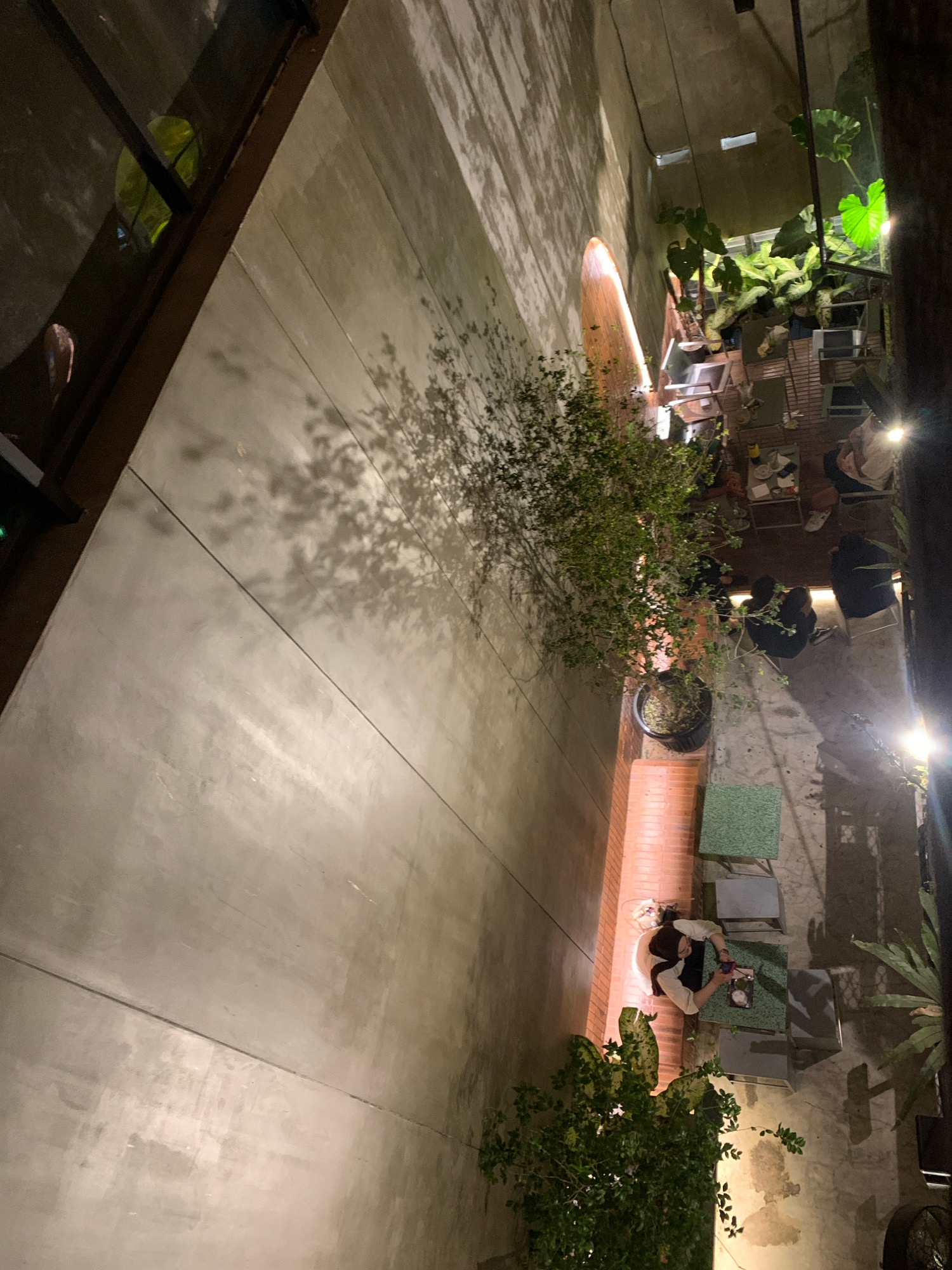 This image, captured at night, depicts an outdoor seating area, possibly a cafe or restaurant, with a contemporary design, featuring concrete walls and floors. The setting is illuminated by artificial lighting, casting shadows from trees onto the textured concrete wall. In the foreground, a person is seated at a table, interacting with their phone. Several other tables and chairs are visible, some occupied by other individuals. Lush green plants, housed in pots, add a touch of nature to the urban environment. The overall scene suggests a relaxed atmosphere, suitable for socializing or enjoying a meal outdoors. The presence of these elements implies that the location is a public establishment that caters to customers seeking an outdoor dining or lounge experience.