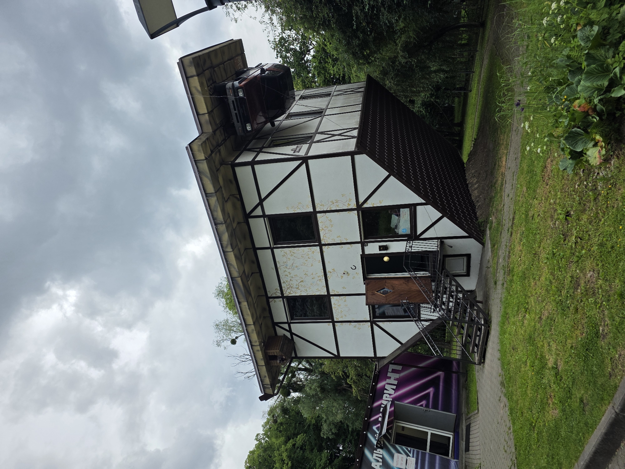 The image shows an upside-down house with a car suspended from its roof, located at the "House Upside Down" attraction in Kaliningrad, Russia. The building has a white facade with dark timber framing and decorative floral patterns. A metal staircase leads to the entrance. Adjacent to the house, a building with a purple and white sign that reads "ЛАБИРИНТ" (Labyrinth) is partially visible. The surrounding environment consists of green grass, trees, and a cloudy sky.