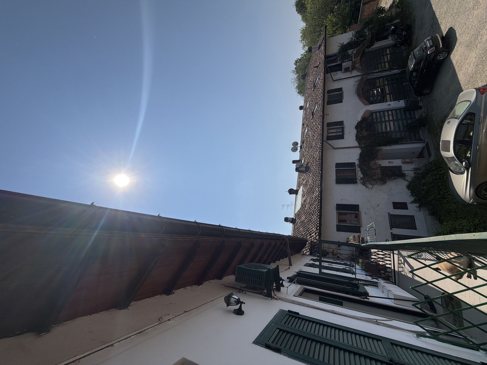 A low-angle shot of a street scene in Milan, Italy, depicts two buildings in close proximity under a bright blue sky with the sun shining. The building on the right is a multi-story white structure with a tiled roof. It features multiple windows with closed shutters, several satellite dishes, and climbing greenery. Two cars and a motorcycle are parked alongside the building. The building on the left is also white, with closed green shutters on the windows, a brown roof overhang, an air conditioning unit, and a light fixture. The viewpoint is from a balcony with a green metal railing, and a cat is visible on the balcony.