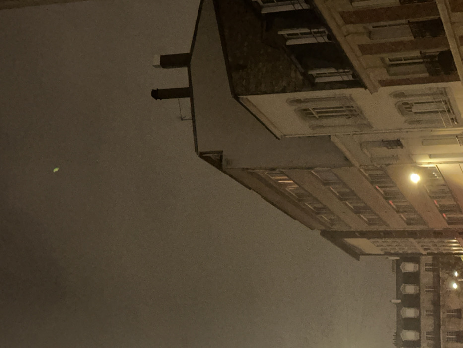 The image depicts a nighttime urban scene in Paris, France, dominated by multiple multi-story buildings under a dark, overcast sky. In the upper-central portion of the sky, a distinct, luminous, green, elongated blur or artifact is visible.
From right to left, the architecture includes:
* A building on the far right featuring a dark, sloped mansard roof with white-shuttered dormer windows and a light-colored facade with decorative window frames.
* Adjacent to it, a light-colored building with several stories, characterized by arched windows on lower floors and rectangular windows above.
* Centrally, a taller, more modern multi-story building with a flat, light-toned facade and rows of rectangular windows, several of which emit a warm orange-red light from within. Its roofline features two prominent, dark-colored chimneys and a small antenna.
* In the mid-ground left, a classic Haussmannian-style building with a grey mansard roof section punctuated by multiple symmetrical dormer windows.
The scene is illuminated by artificial light sources, including a visible street lamp emitting a yellow-orange glow in the lower-left quadrant, and interior lights from various building windows. No people, vehicles, or overt activities are depicted.