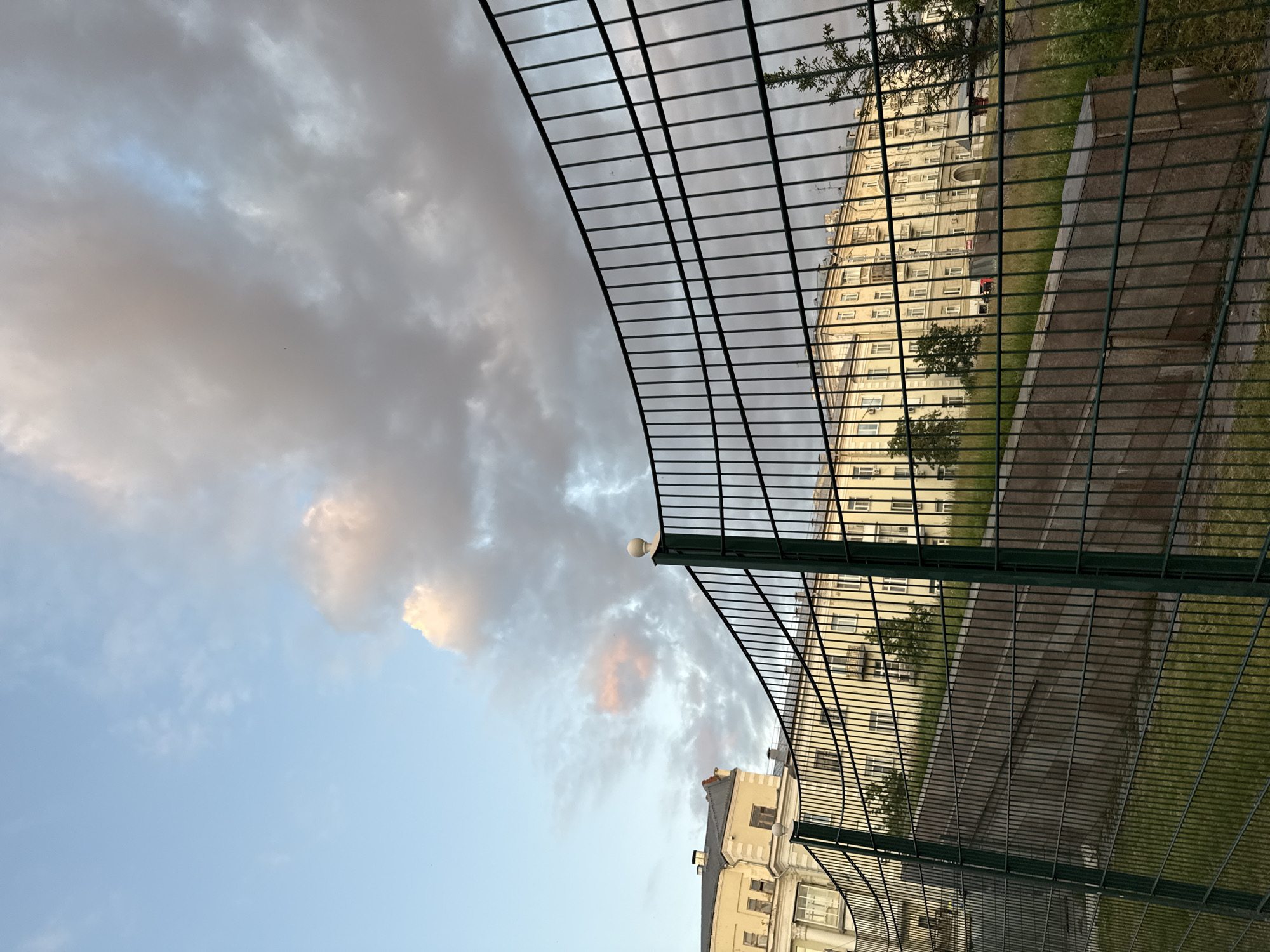 The image shows a view from behind a dark green metal fence, with a curved section in the foreground. Beyond the fence, a building with a pale yellow facade and multiple windows is visible. Some of the windows have balconies. The building appears to be several stories high and has a classical architectural style. In front of the building, there is a sloped area with grassy vegetation and a stone retaining wall. The sky above is partly cloudy, with some patches of light blue sky and darker gray clouds. Some of the clouds are tinged with pink and orange, suggesting it might be sunrise or sunset.
While the image is visually rich, it does not contain enough specific details to pinpoint the exact location within Kazan, Russia. However, the architectural style of the building and the presence of common urban elements like fences and apartment buildings suggest a residential or mixed-use area within a city.