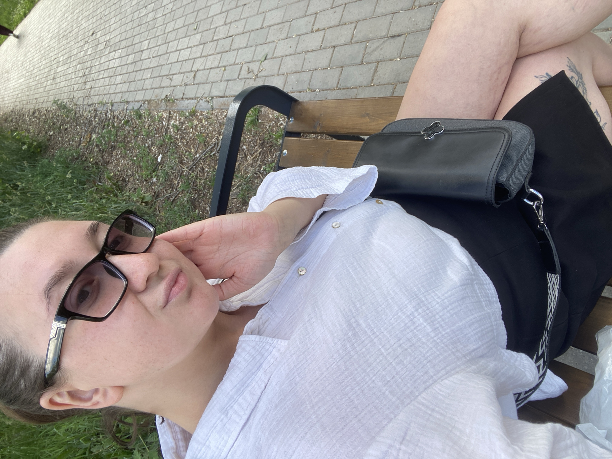 A woman with brown hair pulled back is sitting on a wooden bench in a city park. She is wearing a white, crinkled button-down shirt and a black skirt. She has on black-rimmed glasses and is resting her right hand on her cheek. A black purse with a silver buckle is on her lap. To her right is a paved walkway with a grassy area to the left. The scene appears to be in Chaikovsky, Russia.