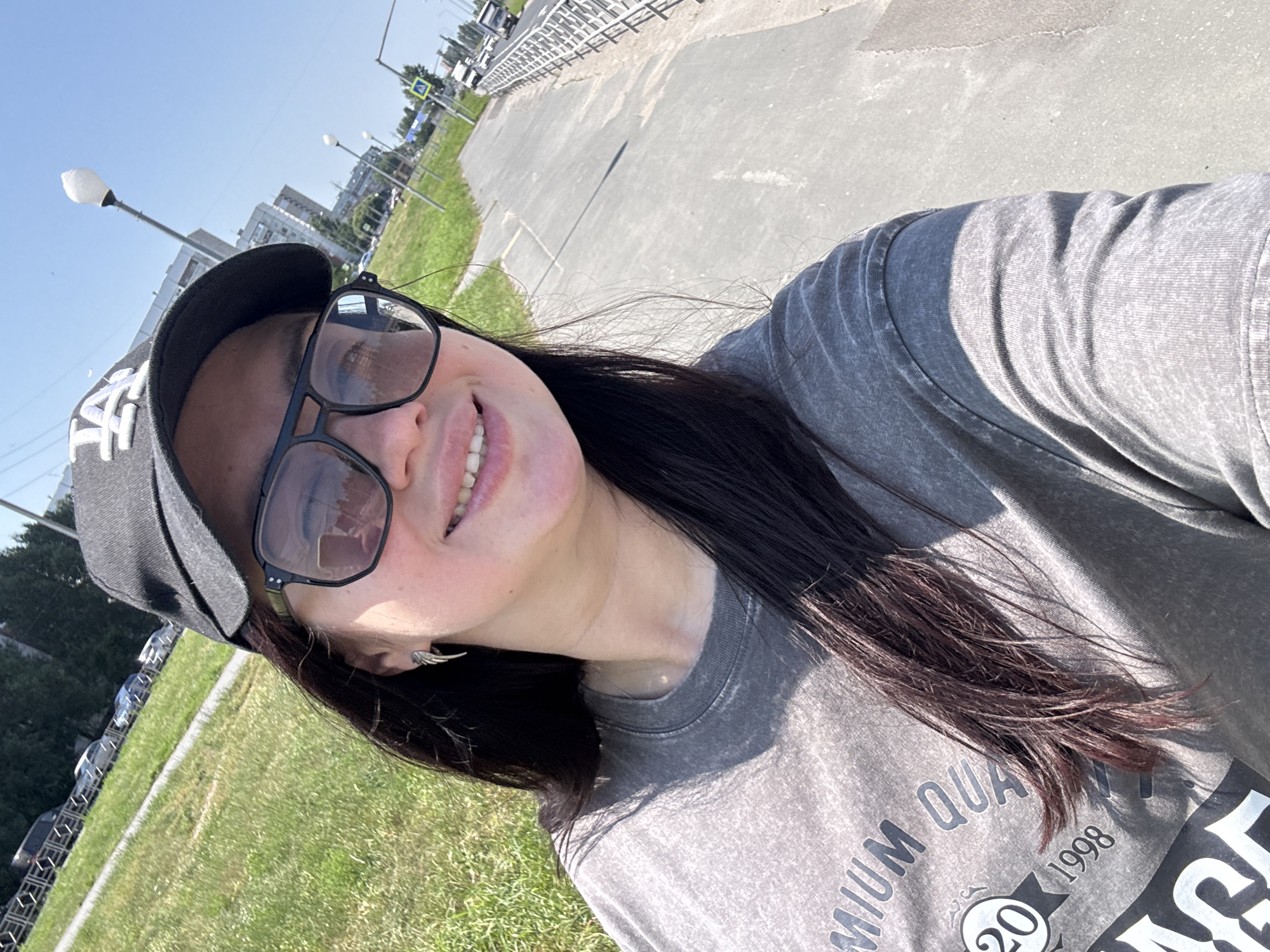 A young woman, wearing a black baseball cap and dark sunglasses, smiles at the camera in a selfie. She has long, dark hair and is wearing a grey t-shirt with text and graphics on it. Her earring is visible, a small silver wing shape. The background shows a sidewalk and a grassy area, with buildings and cars visible in the distance. This scene is likely in the city of Tolyatti, Russia, a city known for its automotive industry and located in the Samara Oblast. The sunlight suggests a clear day.