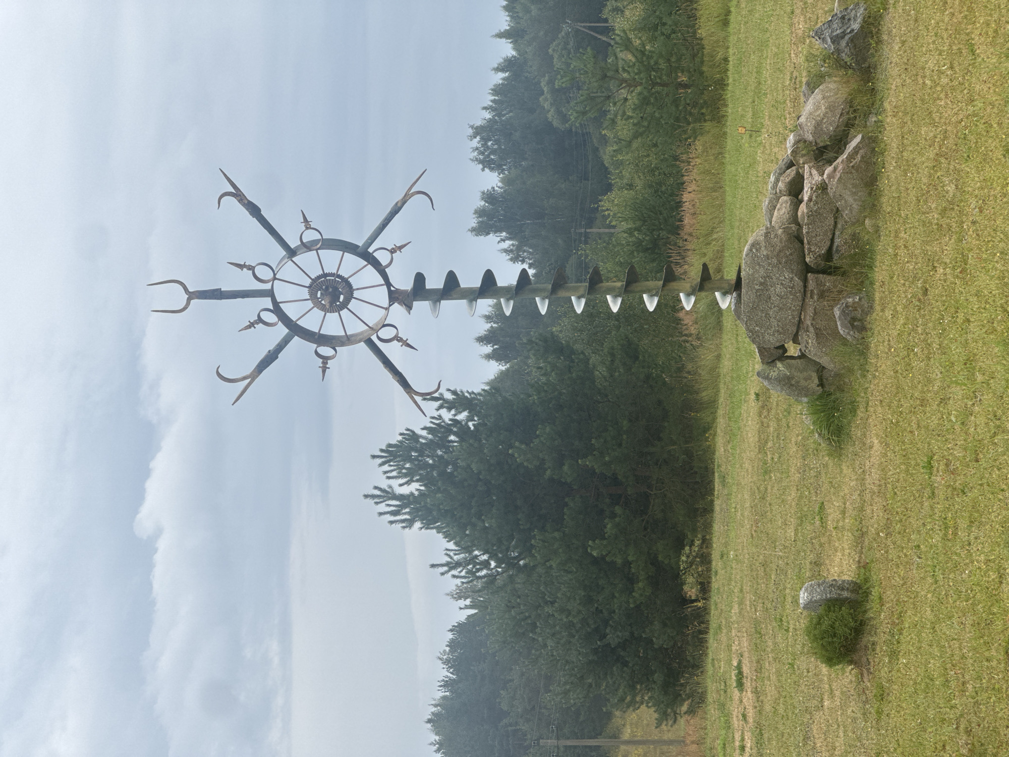 An outdoor, eye-level, full shot captures an abstract metal sculpture resembling a drill bit and a wagon wheel, positioned on a grassy field. The sculpture is anchored by a collection of large, grey stones. Behind the sculpture, a dense forest of pine and mixed trees forms a backdrop under a cloudy, overcast sky. In the foreground, the grass is a mix of green and dry patches, with a small, round stone and a tuft of green shrubbery to the left of the sculpture. A power line pole is visible in the distance to the left of the trees, and a small yellow sign is barely visible in the middle ground to the right of the sculpture. The overall atmosphere is serene and slightly somber due to the cloudy weather.