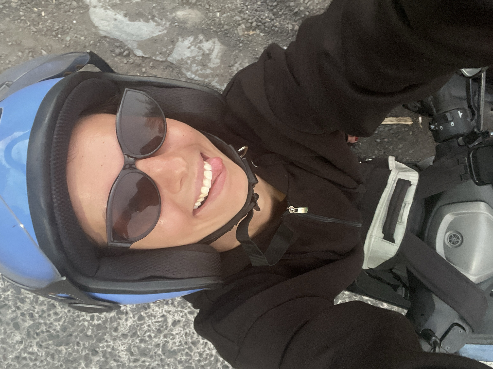 A person is riding a motorcycle in Loloan, Indonesia, and sticking their tongue out at the camera. The person is wearing a blue helmet, sunglasses, and a black hoodie. The motorcycle is black and silver. The background is a paved road with gravel and dirt.