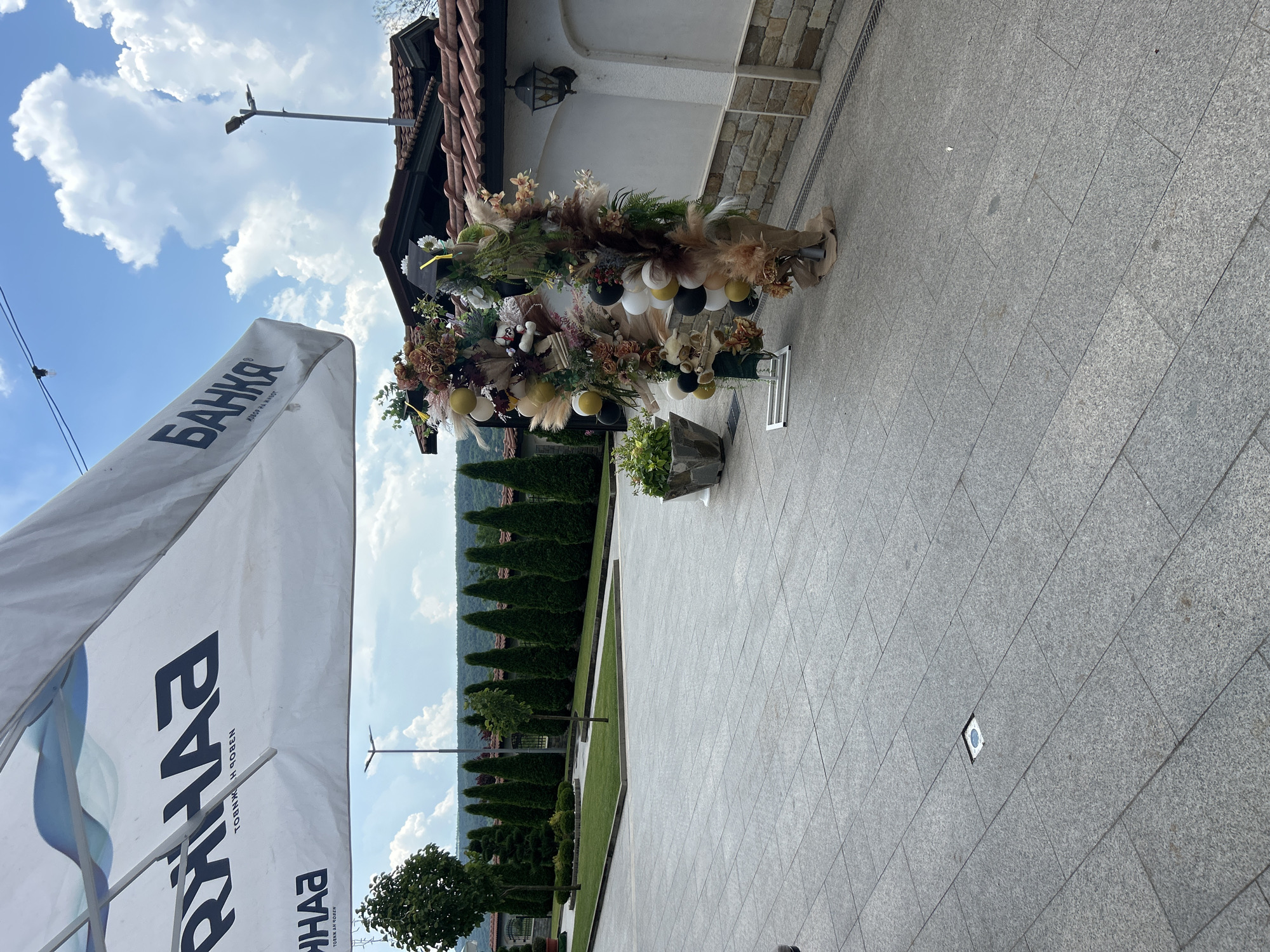 An exterior view of a tiled patio area in front of a building with a red tile roof under a partly cloudy sky. A large white umbrella with the logo "BANJA" in black lettering is partially visible in the upper left corner. To the right of the patio, two tall floral and balloon decorations stand on metal bases. The left decoration is primarily in shades of brown and gold, with some white and black balloons. The right decoration features more green elements, including leafy branches and pampas grass, along with black and white balloons and a graduation cap. A stone wall with a black lantern is visible on the right side of the image. In the background, a row of manicured evergreen trees lines a grassy area. The overall scene suggests an outdoor venue, possibly a restaurant or event space, located in the city of Ruska Bela, Bulgaria.