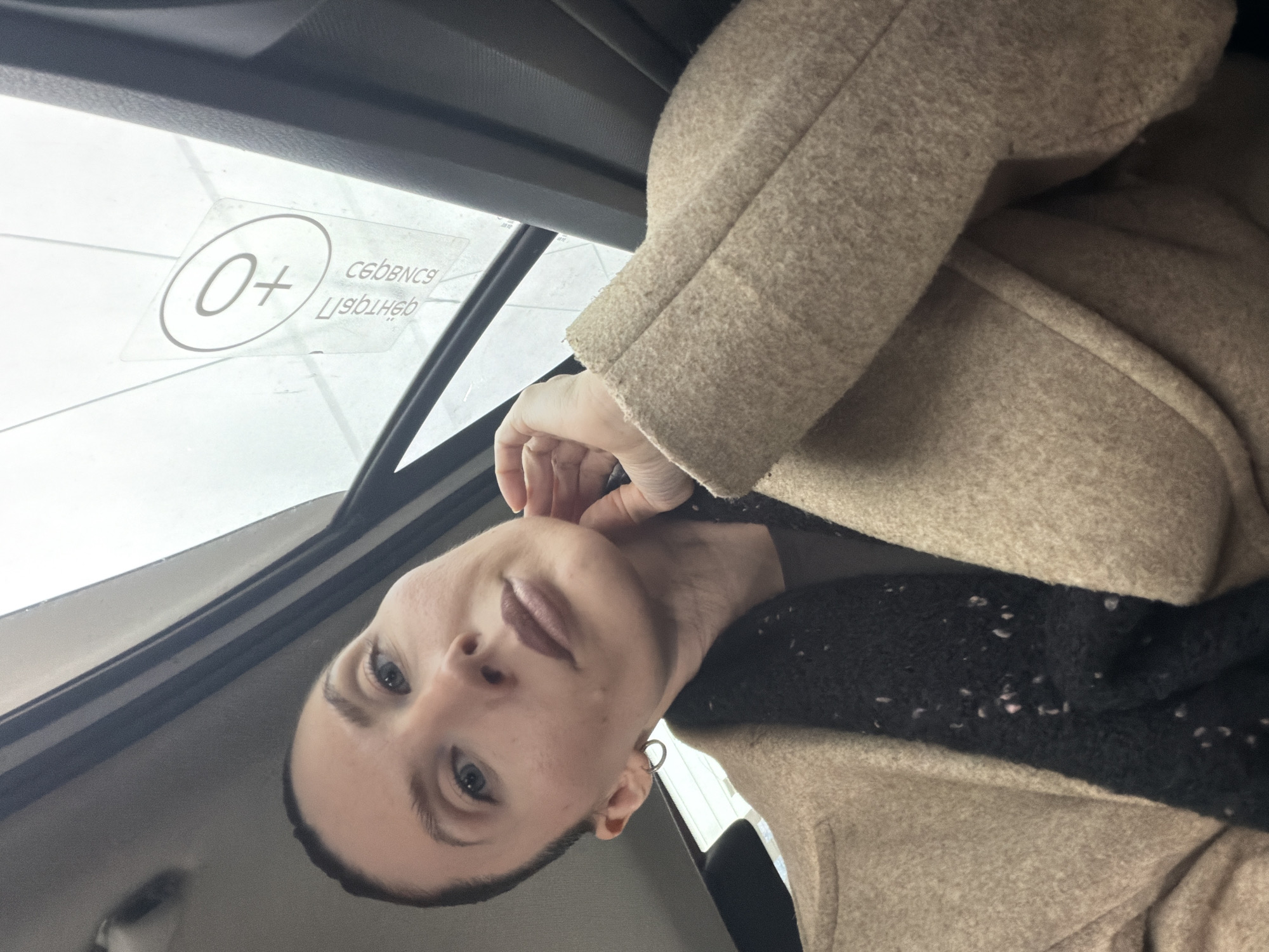 A woman with light skin, short dark hair, and blue eyes is in a vehicle, possibly a car. She is wearing a tan coat and a black sweater. Her left hand is touching her neck. The window shows a "0+" sticker. This indicates the location as Perm, Russia, where such stickers are used to indicate suitability for children.