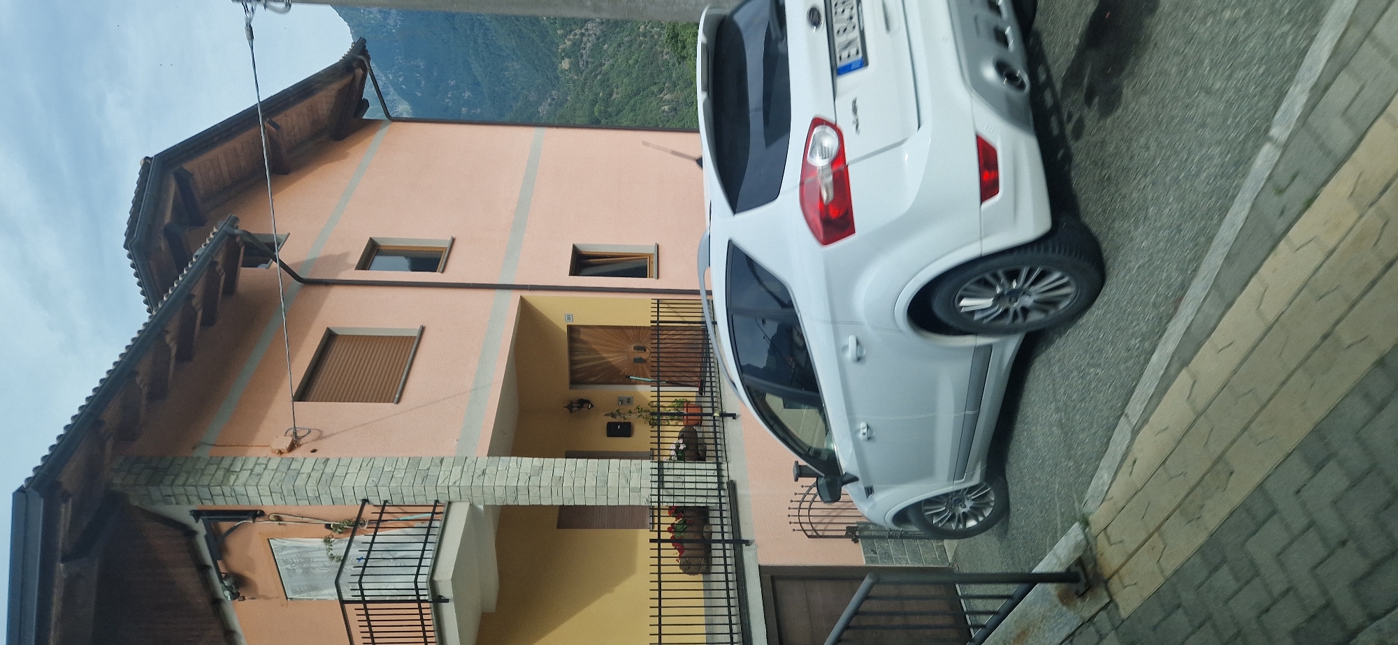 This is a rotated, medium-angle shot of a white SUV parked on a paved street in front of a peach-colored building. The SUV is positioned at an angle, with its rear facing the viewer and its right side closest to the building. The building has multiple windows, some with brown shutters, and a section with yellow siding. A metal fence runs along the front of the building. In the background, a lush green mountainside is visible under a bright sky. The time appears to be daytime, with clear weather. The license plate of the SUV is partially visible and reads "EN 645RP". The scene suggests a residential area in a mountainous region, likely Challand-Saint-Victor, Italy, based on the provided location context. No people are visible in the image.