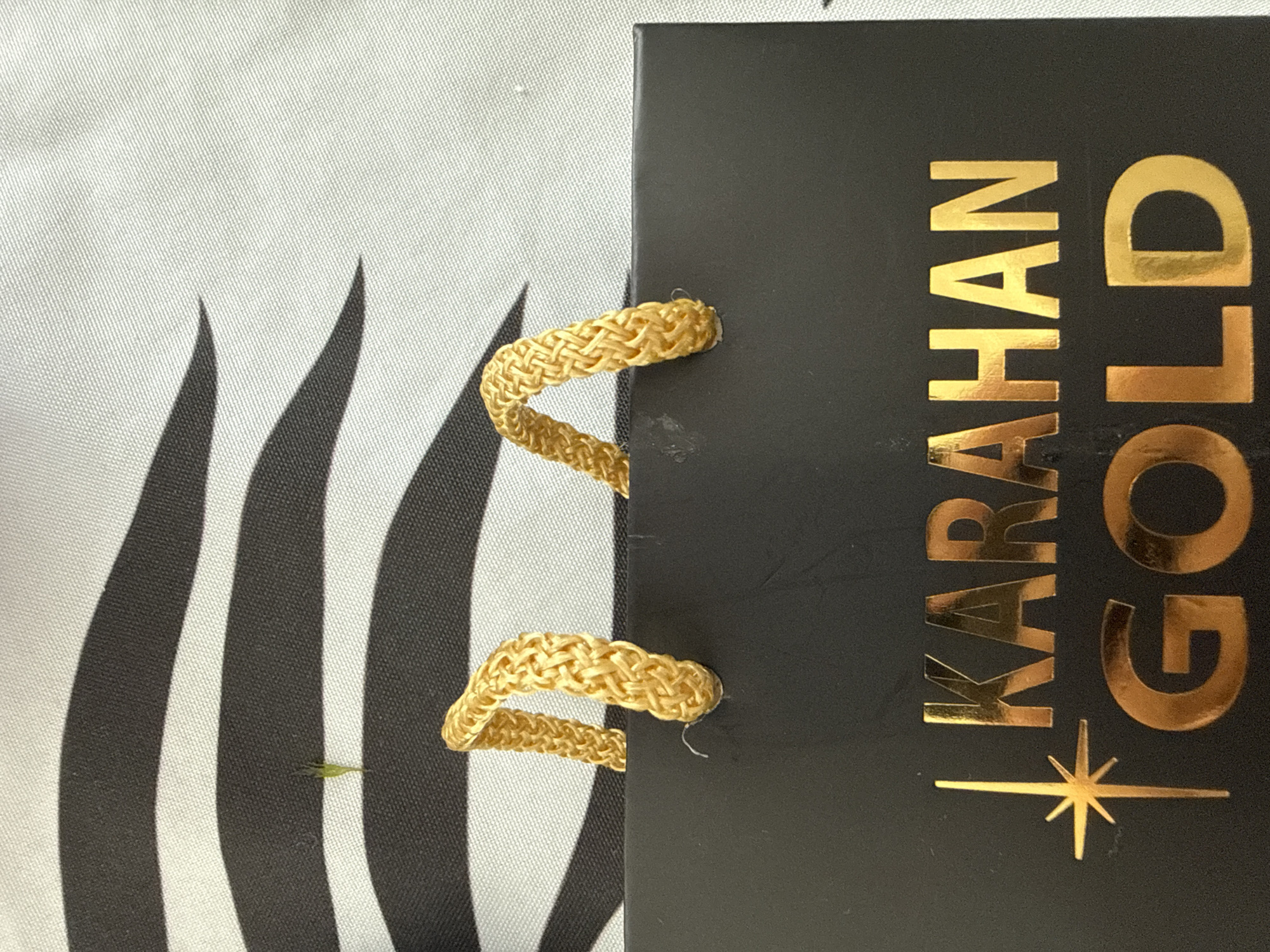 The image shows a close-up of a black gift bag with gold lettering that reads "KARAHAN GOLD". The bag has gold braided handles. In the background, there is a black and white striped pattern. There is a small sprig of green grass on the striped background. The image does not provide any information about the location in the city of Burgas, Bulgaria.