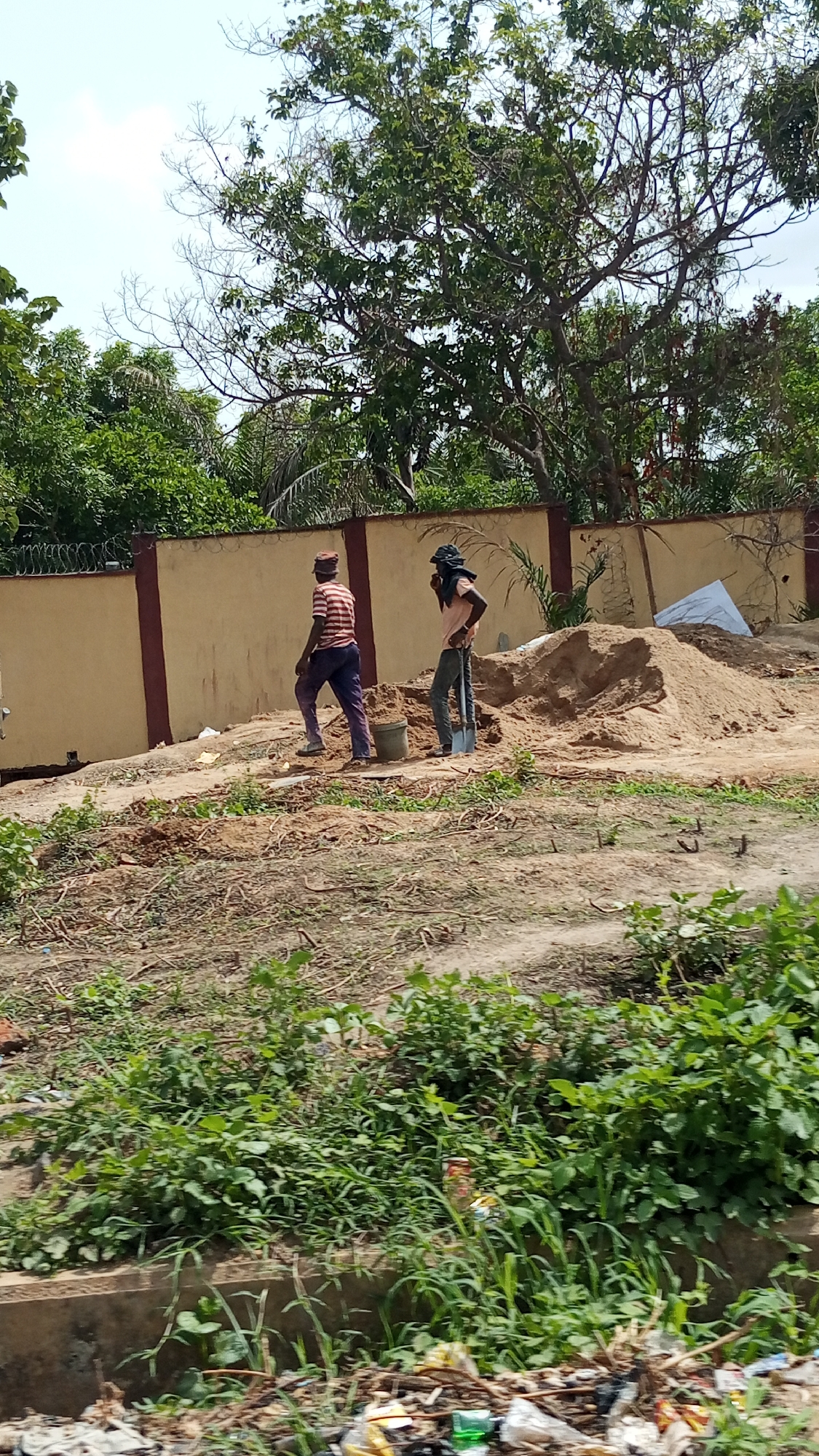 The image depicts an outdoor scene in Zango-ndaji, Nigeria, showing two men engaged in construction or landscaping work. One man, wearing a striped shirt and dark pants, is walking away from a bucket. The second man, with a dark covering on his head and a light-colored shirt, is standing near a pile of sand with a shovel. Behind them is a beige wall with dark brown trim and wire fencing on top. Trees are visible beyond the wall. The foreground shows vegetation and litter. The sky is visible through the trees, suggesting a daytime setting.