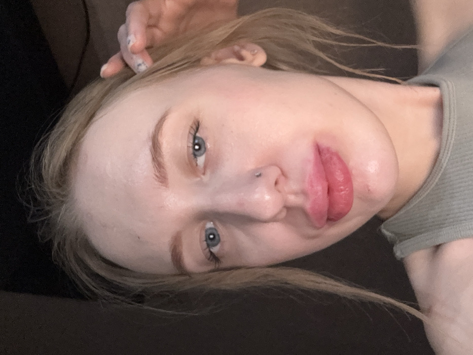 The image presents a close-up portrait of an individual, identified as a young adult female, characterized by fair skin, light blonde hair, and blue irises. A small, metallic stud piercing is visible on the subject's right nostril. The lips are prominent, appearing full and reddish-pink, consistent with recent augmentation or swelling. The individual is wearing a light olive-green ribbed tank top. A partial view of the subject's right hand, displaying light-colored fingernails, is present in the upper right quadrant, seemingly touching the hair near the right temple. The background is uniformly dark and out of focus, providing no discernible environmental details or objects. The specific location within the city of Rostov-on-Don, Russia, cannot be determined from the visual data provided in this image.