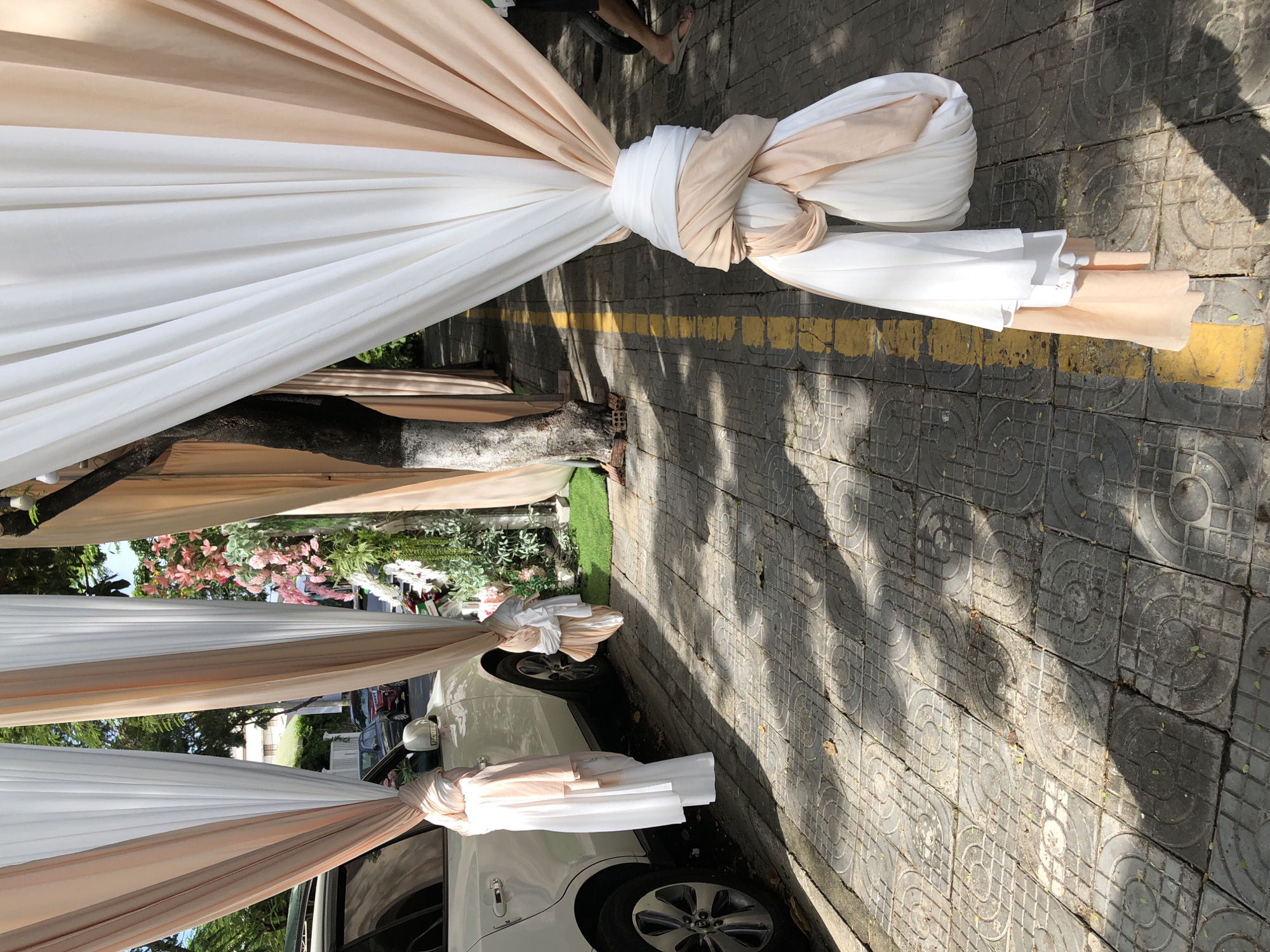 This is a photograph of a pathway decorated with drapes of white and peach fabric. The pathway is paved with square tiles, some of which have a circular pattern. A yellow line runs down the center of the pathway. To the left, a white SUV is parked, adorned with similar drapes tied to its side mirrors and door handles. Beyond the SUV, foliage and a building are visible. To the right, a tree stands with its trunk partially wrapped in fabric. Further down the pathway, a person's legs and feet, wearing flip-flops, are visible. The scene suggests an outdoor event, possibly a wedding or festival, taking place in an urban environment, likely in Da Nang, Vietnam, given the architectural style of the building in the background and the presence of palm trees, which are common in the region. The lighting indicates it is daytime, with shadows cast on the ground from the drapes and foliage.