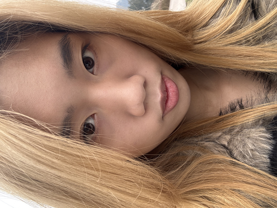 A close-up shot of a young woman with light blonde hair framing her face. Her skin appears fair with a subtle pink undertone on her lips. She gazes directly at the camera with soft brown eyes. Her eyebrows are dark and well-defined. She wears what looks to be a jacket with a furry collar, partially visible on the right side of the frame. The image is slightly rotated, adding a dynamic angle to the composition.