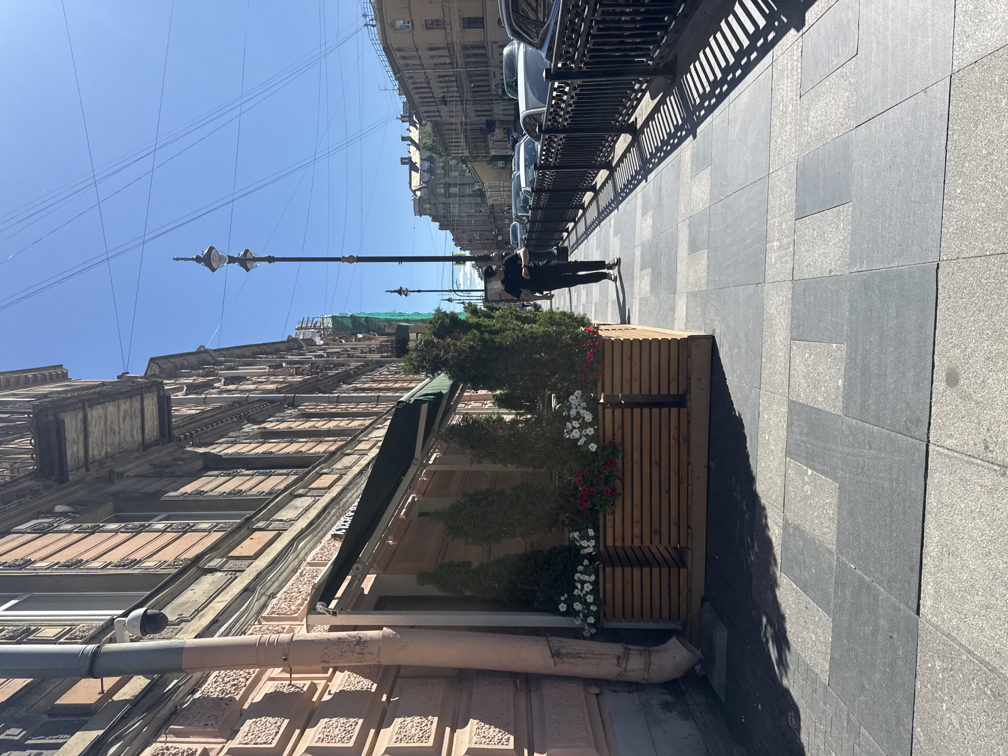 The image shows a street scene in Saint Petersburg, Russia on a sunny day. The perspective is from a low angle, looking up at a building and the sky, with the sidewalk and street to the right.
In the foreground, a light pink building with intricate architectural details and visible pipes runs along the left side of the frame. Further down the street, a modern wooden bench with integrated planters filled with red and white flowers and greenery stands on the sidewalk. A person in dark clothing, wearing a baseball cap, stands near the bench, casting a shadow on the pavement.
The sidewalk is paved with large, dark grey tiles interspersed with lighter grey granite tiles, forming a geometric pattern. A black metal fence runs along the edge of the sidewalk, separating it from a row of parked cars. The buildings across the street are old, multi-story structures with classic European architecture. Overhead, a street lamp with two bulbs is visible against a clear blue sky crisscrossed by numerous thin wires. The sunlight suggests it is daytime, likely late morning or early afternoon, given the sharp shadows. The overall mood is one of urban tranquility on a bright, clear day. No text is visible in the image.