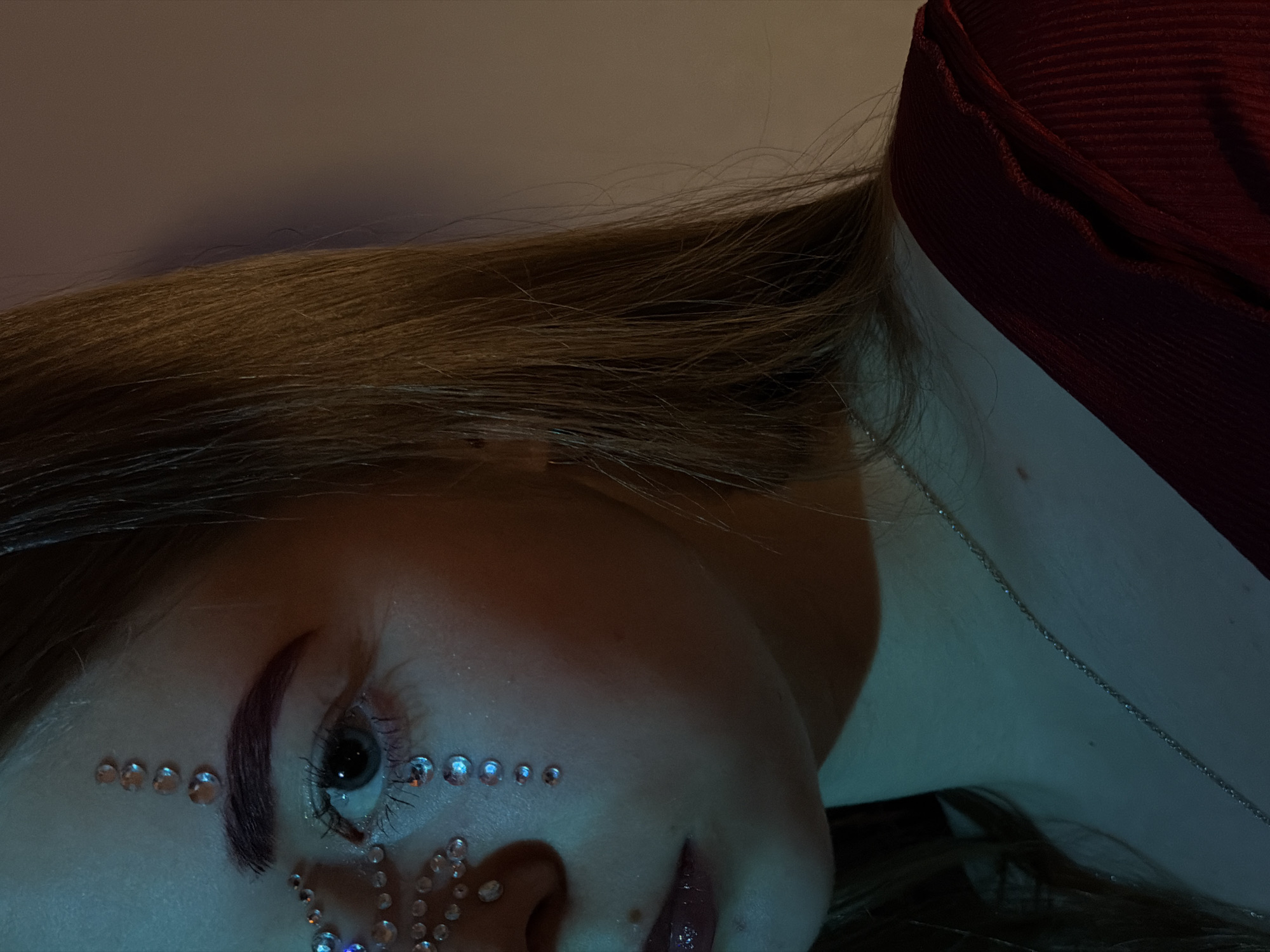 The image shows a close-up of a person's face and neck, angled towards the left. The person has fair skin, brown hair, and is wearing makeup with small rhinestones arranged around the eye and eyebrow. A silver chain necklace is visible on the neck. The background is a gradient of light colors, with a possible red garment visible in the upper right. The lighting is muted and appears to be artificially produced, casting a cool hue on the scene. There is no information to indicate the location is in Perm, Russia.