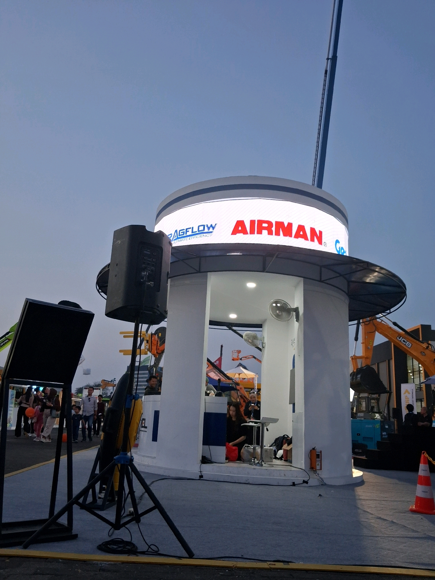 A tall, cylindrical structure with a curved roof is the central focus of the image, illuminated by a large, circular LED display at its top that prominently features the word "AIRMAN" in red. Beneath the display, the structure is open, revealing a white interior with a circular bar or counter. Inside, a woman in a black dress is seated on a stool, and a small table with a red bag on it is nearby. Two wall-mounted fans are visible on the interior walls. A large black speaker is mounted on a tripod to the left of the structure. In the background, to the right, an orange construction vehicle, identified by a "JCB" logo, is parked. A blue generator is positioned in front of it. People are visible in the background, appearing to be at an outdoor event or exhibition. The sky is a clear, light blue, suggesting it is either early morning or late afternoon. The scene appears to be an outdoor exhibition space, possibly at JIExpo Kemayoran in Central Jakarta, Indonesia.