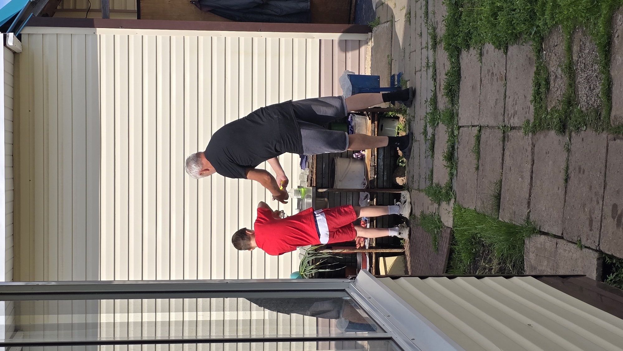 The image depicts an outdoor scene in Забегалово, Russia, featuring an adult male and a child male engaged in an activity at a rustic outdoor sink. The adult, with grey hair, wears a black t-shirt, grey shorts, black socks, and dark slip-on shoes; he stands on the right, facing left, with his hands positioned over the sink, holding a yellow sponge. The child, wearing a red t-shirt, red shorts over a white waistband, white socks, and light-colored sneakers, stands on the left, facing right, also with his hands in the sink basin.
The sink is integrated into a multi-tiered, utilitarian workstation constructed from rough materials, possibly wood and metal. To the left of the sink, green leafy vegetables are visible on a surface. A white pump-action soap dispenser and folded fabric items are on the workstation to the right of the sink. Various buckets and containers are stored on shelves below the workstation.
The background consists of a building clad in light beige horizontal siding. A large window on the far left reflects portions of the building and the sky. To the far right, an open doorway reveals a dark coat or garment hanging inside. The ground is composed of rectangular concrete pavers forming a pathway, with grass and weeds growing prominently in the gaps between them. The scene is illuminated by direct sunlight, casting distinct shadows.