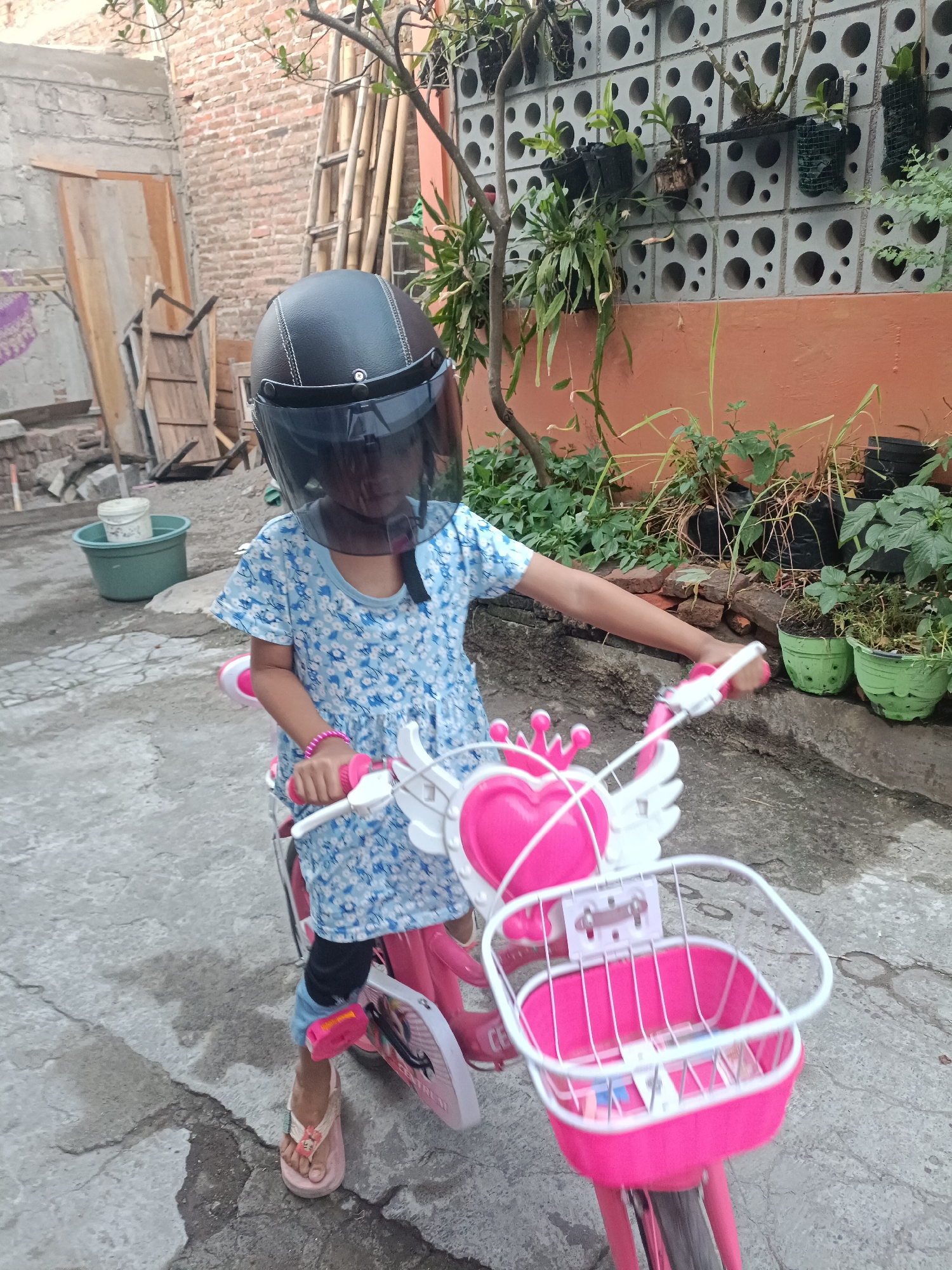 In Jatipuro, Indonesia, a young girl, wearing a blue floral dress, black leggings, and pink flip-flops, poses on a pink bicycle. She is equipped with a black helmet with a transparent visor, obscuring her face. Her hands are on the handlebars of the bicycle, which features a pink heart-shaped accessory with wings and a white basket. The background shows a brick wall, bamboo poles, and potted plants, suggesting an outdoor, possibly residential, setting. The ground is paved with concrete.