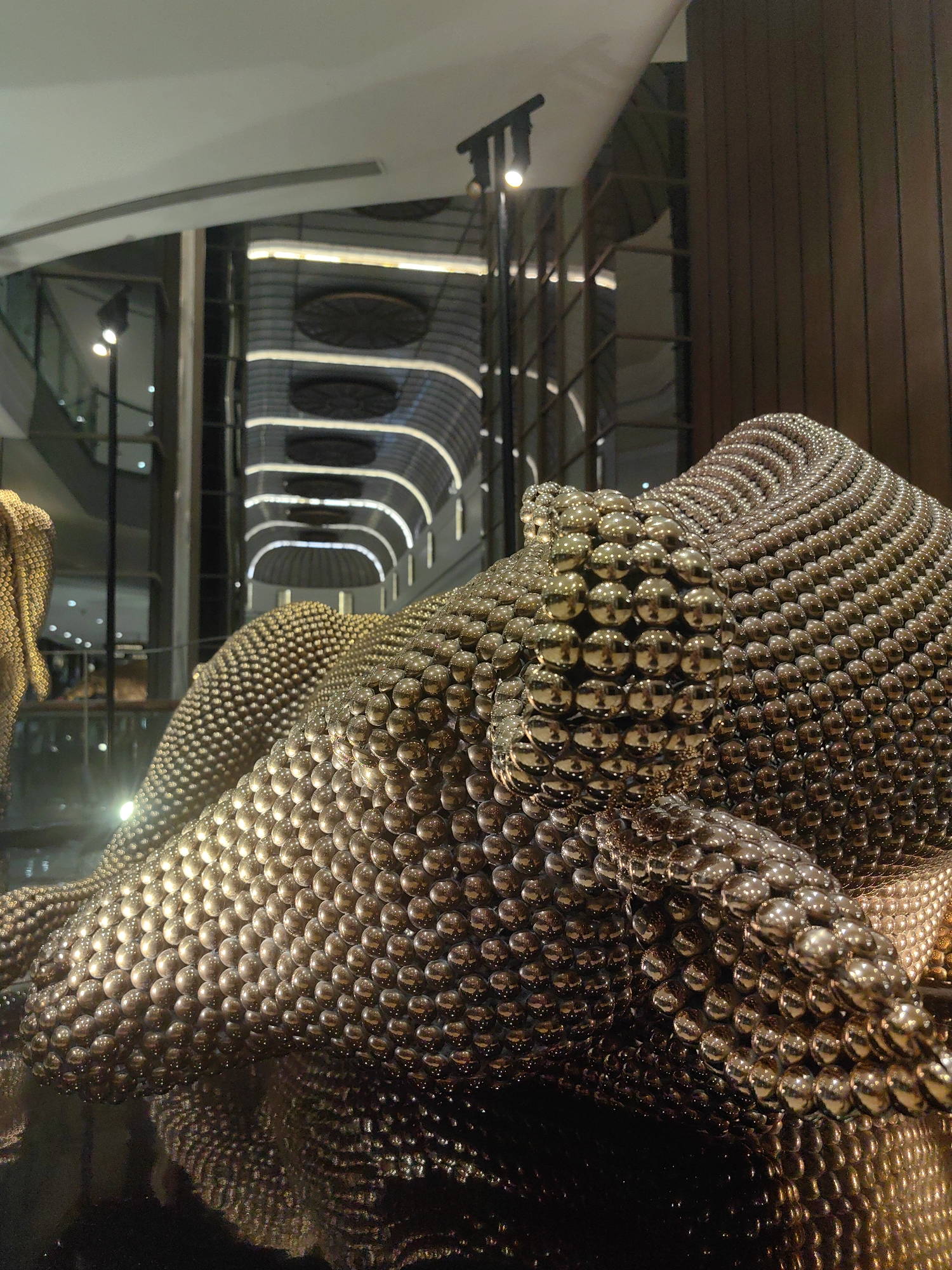 The image shows a close-up view of a metallic sculpture made of small, interconnected spheres. The sculpture appears to be abstract and takes on a flowing, undulating form, possibly representing a draped fabric or a body. The spheres are highly reflective, creating a shimmering effect and casting reflections on the surface below, which also appears to be reflective and dark. In the background, blurred elements suggest an indoor environment with architectural features such as curved ceilings with integrated lighting and vertical wooden panels. The overall impression is one of a contemporary art installation within a modern interior space, possibly a gallery, museum, or upscale commercial establishment. There are no people visible in the image. The style of the sculpture and the background architecture do not provide specific clues to pinpoint the exact location in Mumbai, India, but it could be found in an art gallery or a shopping mall.