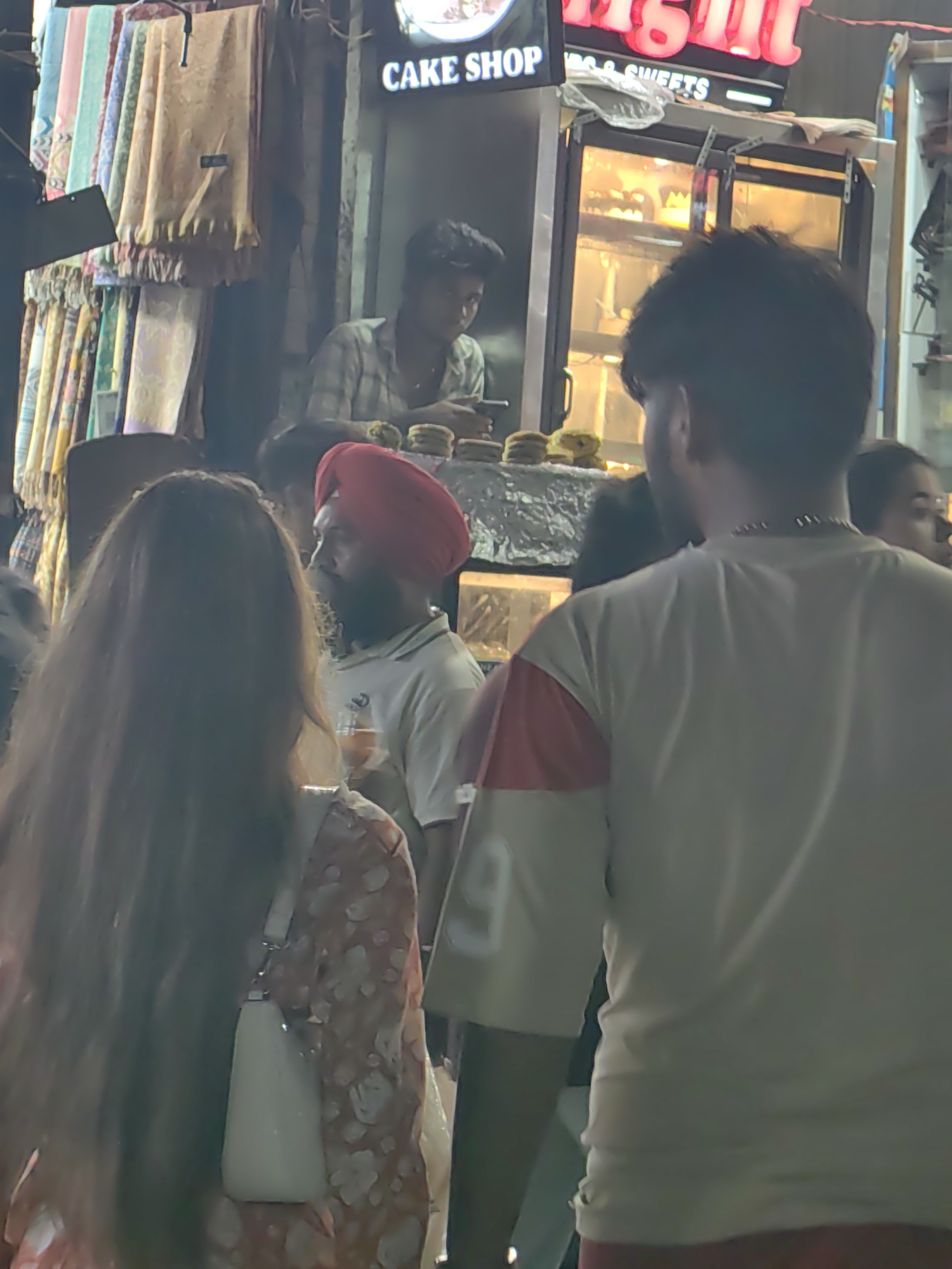 The image shows a street scene in Manali, India, likely a bustling market area. In the foreground, a person with long dark hair is facing away from the camera, wearing a floral-patterned dress and carrying a white purse. To their right, another person is visible from behind, wearing a light-colored t-shirt with red and pink sleeves and a chain necklace.
In the mid-ground, a man wearing a red turban and a light-colored polo shirt is standing near a food stall. He appears to be holding a drink and looking to his right. Behind him, a person in a plaid shirt is visible, leaning over a counter.
The background reveals a "CAKE SHOP" sign with neon lights, and a refrigerated display case filled with baked goods. To the left of the cake shop, there are racks of colorful scarves or textiles hanging for sale. The overall scene suggests an outdoor market or shopping street with food vendors and retail stalls, typical of a tourist destination like Manali.