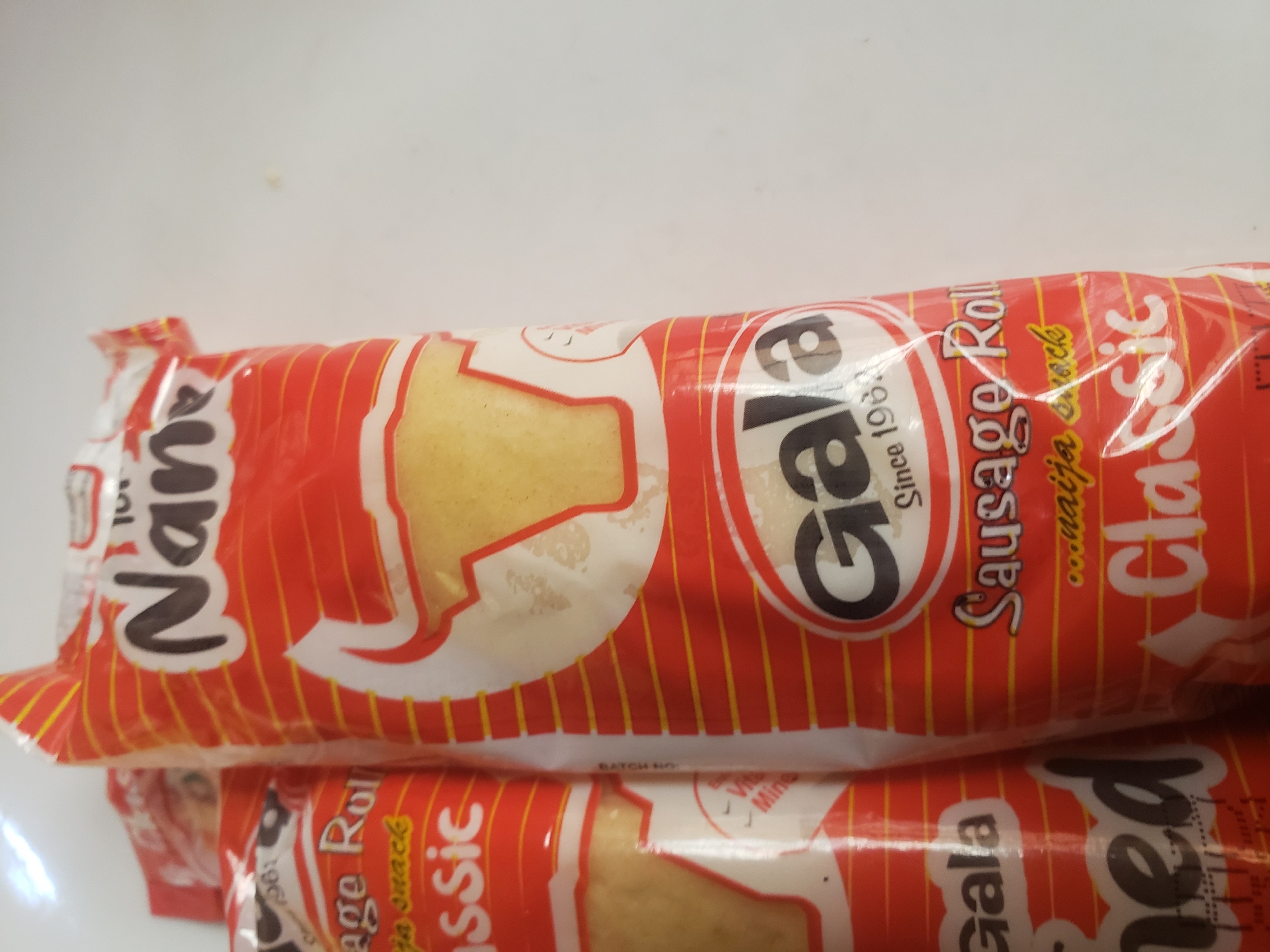 The image shows a close-up of two packaged sausage rolls, identified as "Gala" and "Nana". Both are primarily red with yellow stripes and white lettering. The "Gala" package features a black oval with "Gala" written in white, followed by "Since 1963". Below this is "Sausage Roll" and "...naija snack". Further down, the word "Classic" is visible in white lettering. The "Nana" package, partially visible on the left, also has "Nana" in large black letters. Both items appear to be snacks. The background is a plain white surface, suggesting an indoor setting, likely a shop or pantry. The lighting is somewhat diffused. There are no people or any discernible indication of the specific location within Sabon Tasha, Nigeria, beyond the branding of the products.