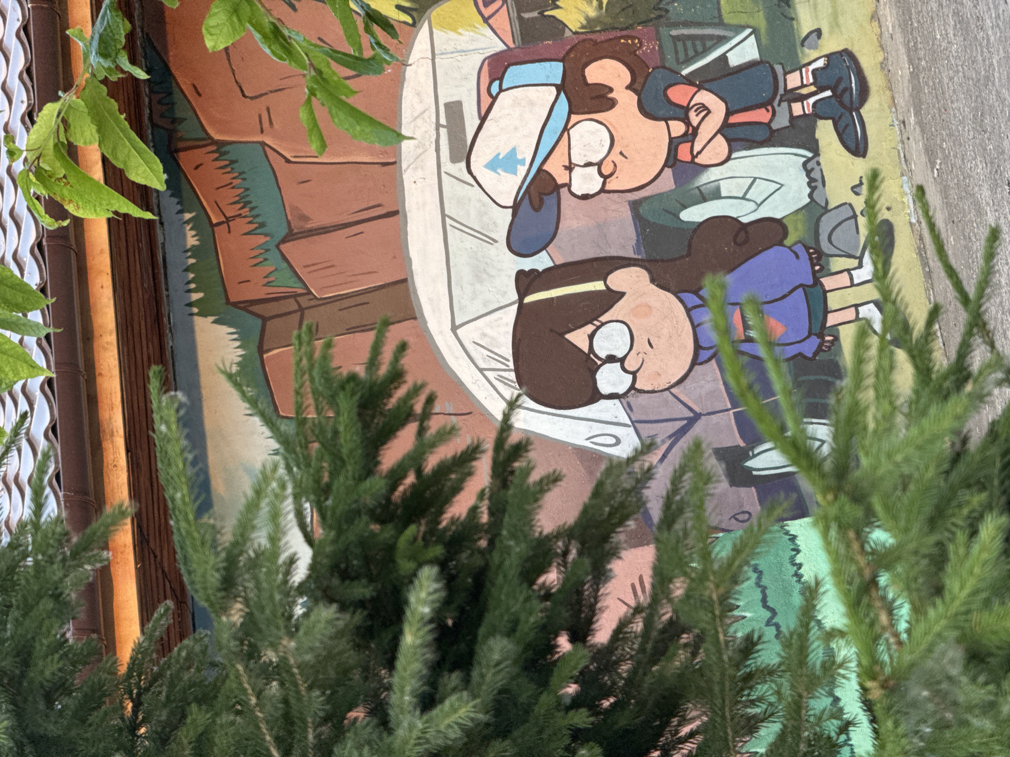 The media file is a photograph of a mural featuring two cartoon characters, likely from the animated series Gravity Falls, based on their appearances. One character is a young boy with brown hair wearing a blue hat with a pine tree symbol, a red shirt, and blue shorts. He is lying down, resting his head on his arm. The other character is a young girl with brown hair wearing glasses and a purple sweater. She is also lying down. The mural is painted on a wall, with elements of nature like trees and rocks depicted. The style is illustrative and colorful.
The photo is taken from an angle, with green foliage from real plants obscuring parts of the mural and the background. The background appears to be an outdoor setting, possibly a street or a courtyard, with a concrete surface visible to the right. The lighting suggests it is daytime. The mural itself depicts a playful or relaxed scene.
No visible text is present in the media file. The location context provided is Krasnoyarsk, Russia.