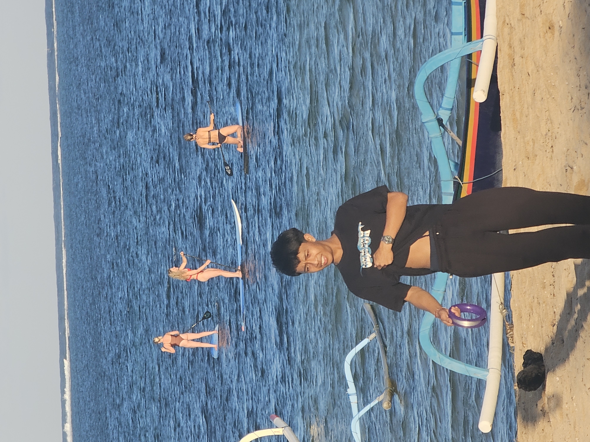 The image depicts a beach scene in Denpasar, Indonesia, likely in the early morning or late afternoon due to the soft lighting. In the foreground, a young man wearing a black t-shirt and black pants stands on a sandy beach. He is holding a purple ring in his left hand and pulling up the hem of his t-shirt with his right hand, revealing his abdomen. He is looking towards the right side of the frame with a neutral expression.
In the background, the blue ocean stretches out. Several people are engaged in stand-up paddleboarding. Three women are visible on paddleboards: one in a brown bikini on the left, another in a red bikini in the middle, and a third in a black bikini on the right, kneeling on her board. The paddleboards are on calm water, with subtle ripples indicating a gentle breeze. In the shallow water near the beach, a traditional Indonesian outrigger boat, painted in dark blue with red, yellow, and black stripes, is partially visible. The boat has outriggers made of white and blue tubes. The sky above is a pale, clear blue.