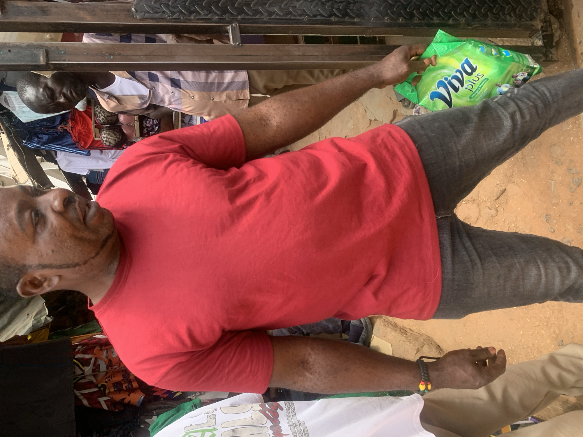 The image shows an adult man standing in what appears to be an outdoor market setting in Jos, Nigeria. He is wearing a red t-shirt and gray trousers, holding a green package of "Viva Plus" detergent. Another man, wearing a patterned shirt, is visible in the background along with various articles of clothing displayed for sale. The ground is dirt or sand. A metal structure, possibly part of a stall, is visible above. The man holding the detergent has some facial markings. A bracelet is on his left wrist.