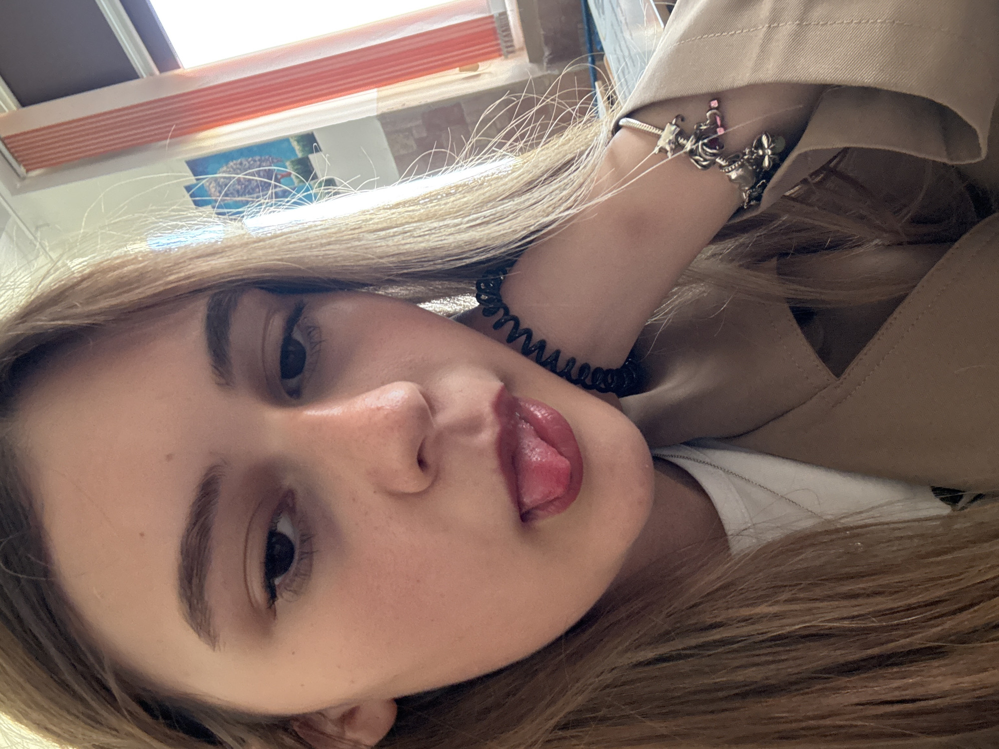 A young woman with long blonde hair is angled slightly, her head tilted to the left. She has fair skin, dark eyes, and slightly parted lips with her tongue visible. Her right hand is raised near her head, adorned with two bracelets: one a silver charm bracelet and the other a black coiled bracelet. She wears a beige jacket and a white shirt underneath. The background includes a window with orange trim and a partially visible picture or artwork. The location of this image is not provided. The city of Panagyurishte, Bulgaria is not mentioned.