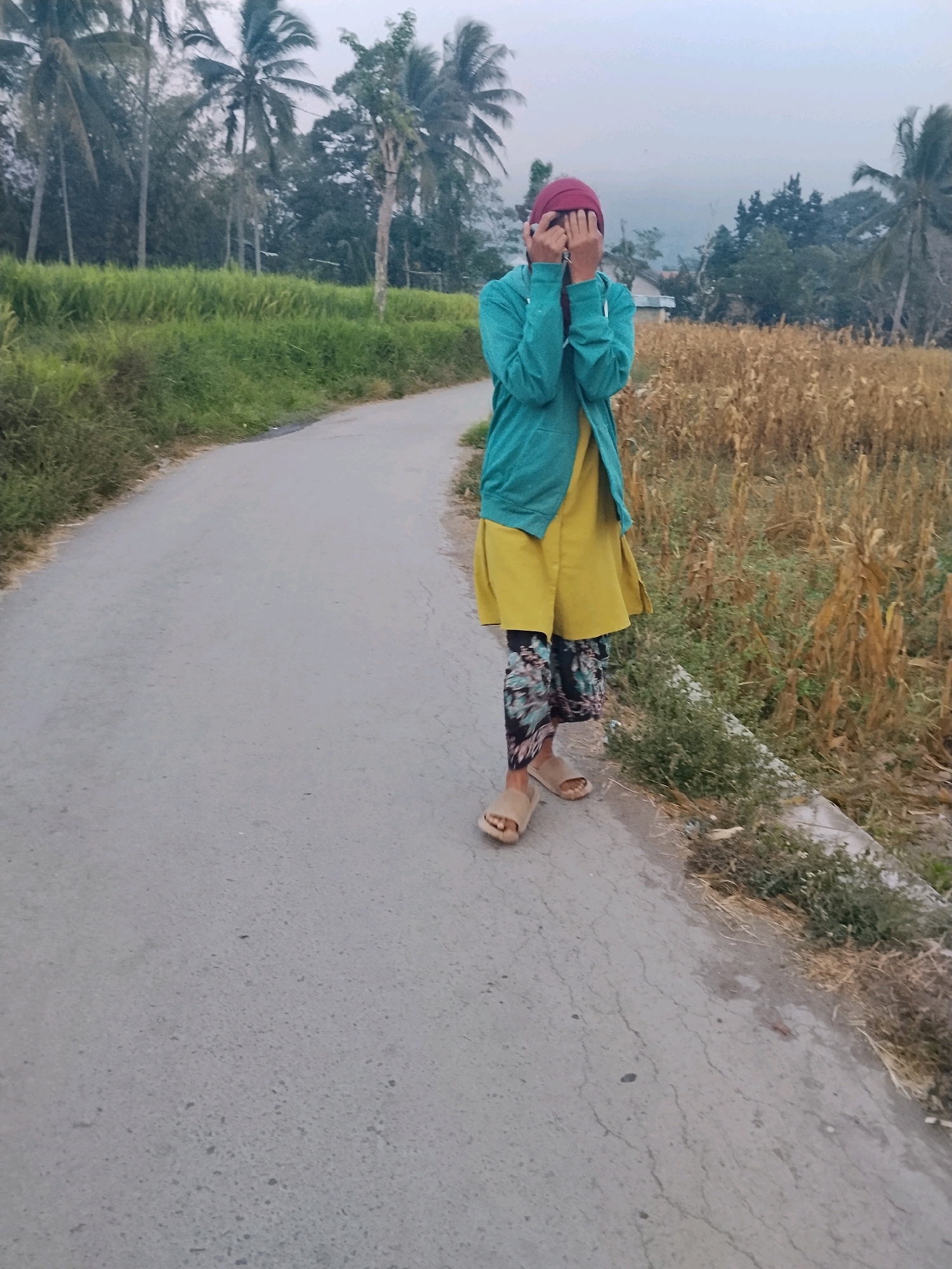 A person stands on a paved road in Tlogosari, Indonesia. The person is wearing a red headscarf, a turquoise jacket over a yellow dress, and patterned leggings. They are covering their face with their hands. To the right of the road is a dry field, likely of corn stalks, and to the left is a lush green area. In the background, coconut trees are visible, indicating a tropical environment. The sky is overcast.